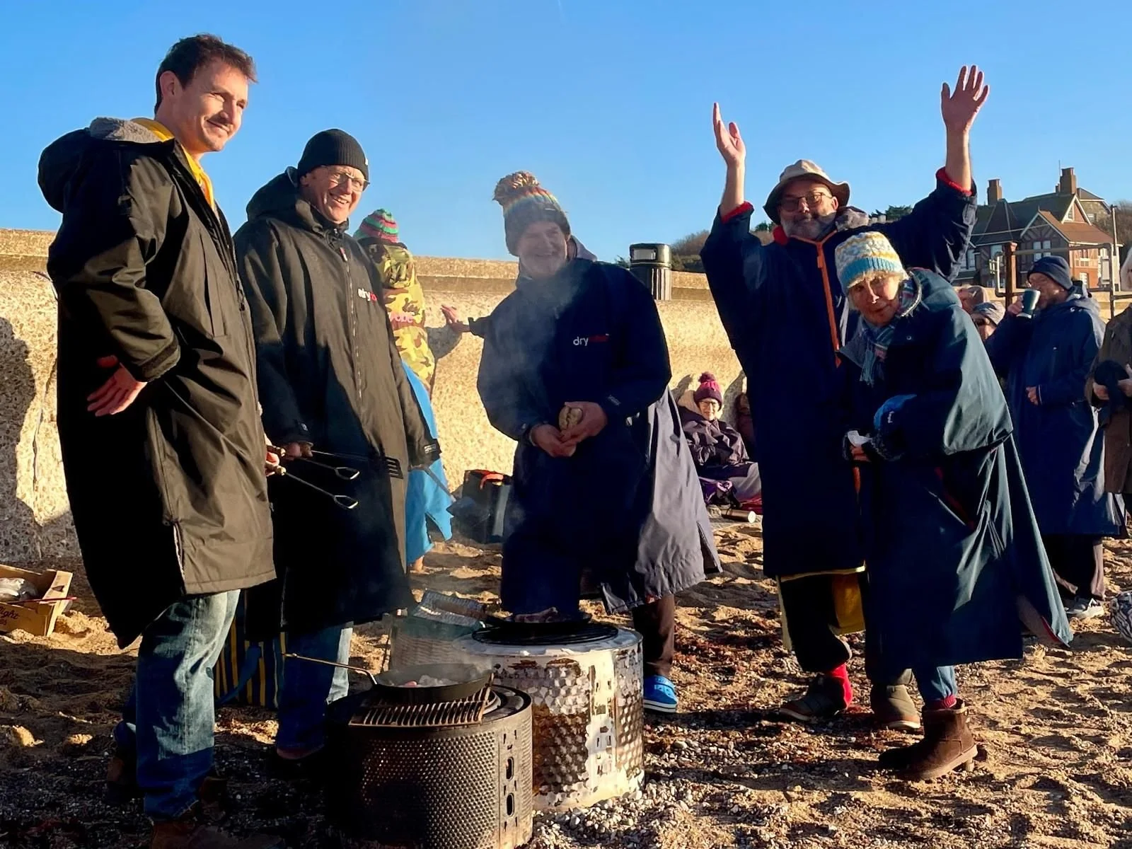 A group of people gathered outdoors on sandy ground, wearing warm clothing and hats, enjoying a social event with some grilling food on portable barbecue grills. The sky is clear and blue.