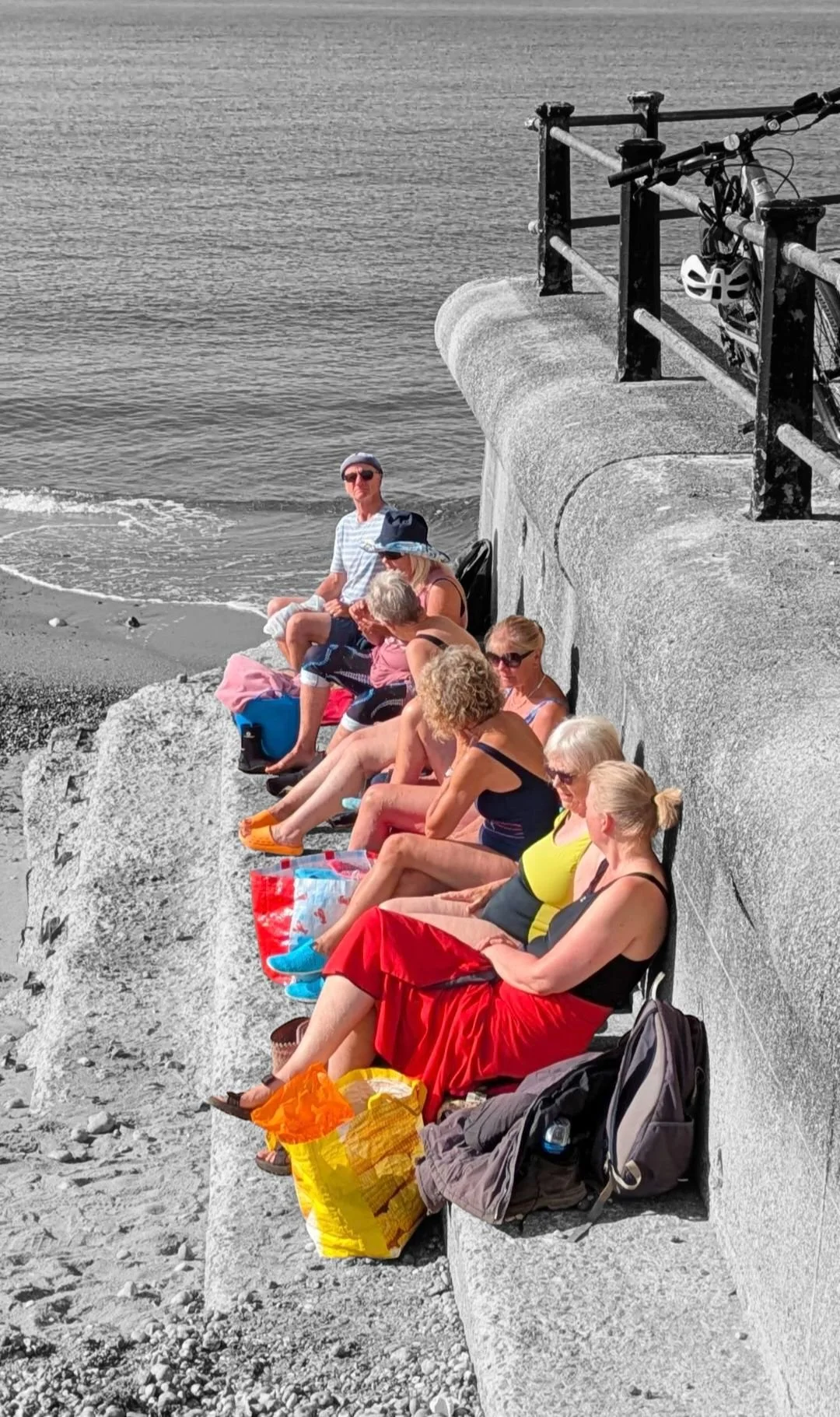 Group of people sitting on a concrete ledge near the water, some with bags and personal belongings, enjoying a sunny day at the beach.