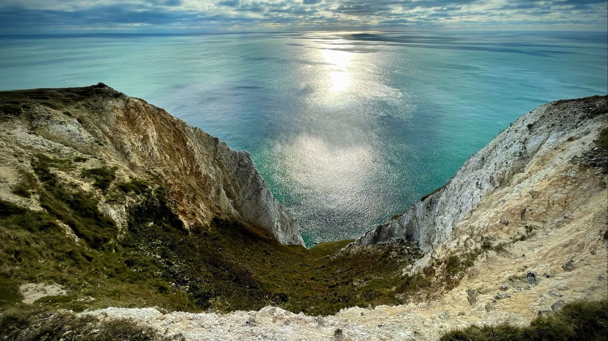 Cliffside view of the ocean with sparkling water and a partly cloudy sky.