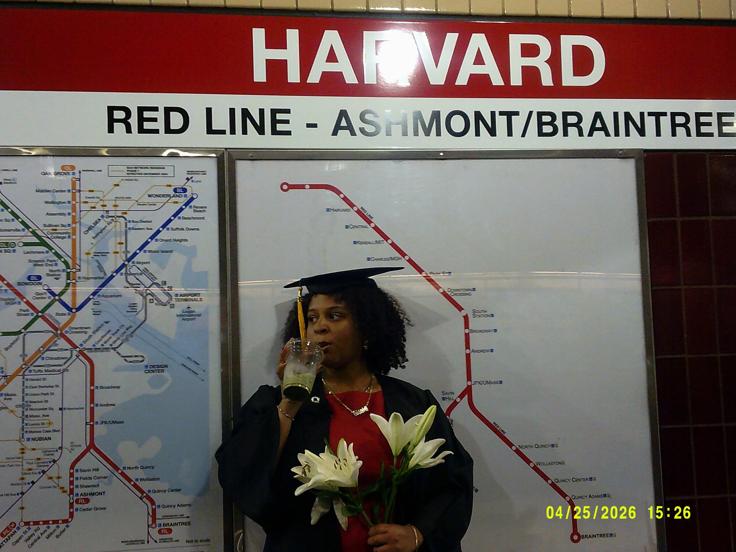 A woman with curly hair in graduation attire holding a bouquet of white lilies and drinking from a clear bottle in front of a subway map and sign indicating Harvard station on the Red Line, Ashmont/Braintree branch.