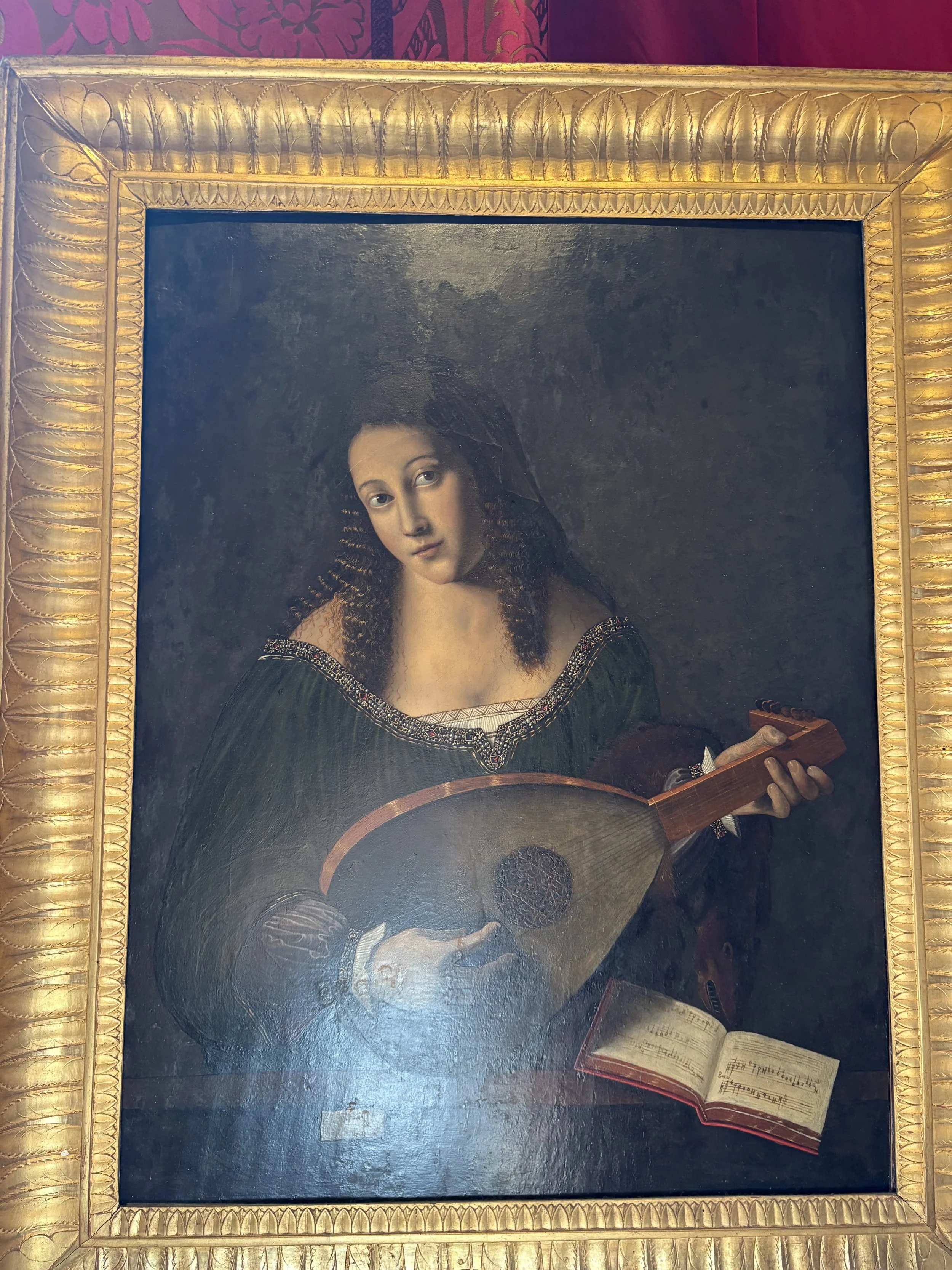 Portrait of a woman with long, curly hair holding a guitar, wearing a dark dress with detailed embroidery, framed with an ornate gold border, against a dark background