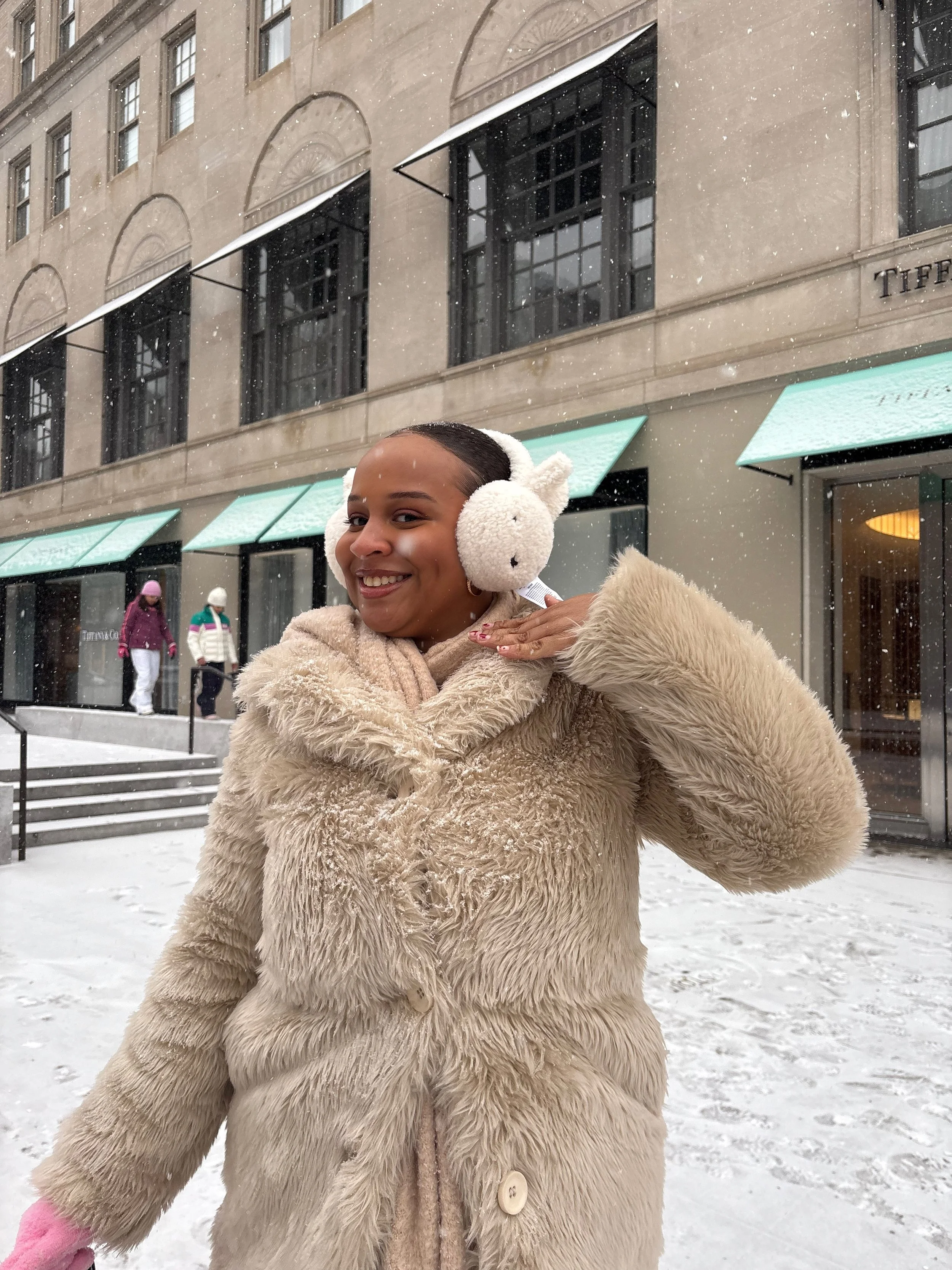 A woman wearing a fluffy beige coat and matching earmuffs smiling outdoors in snowy weather.