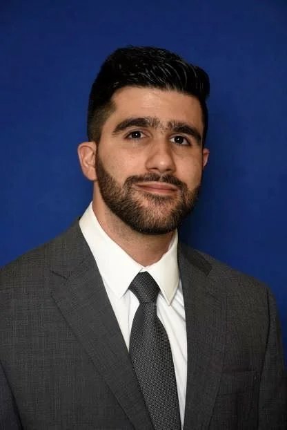 Professional headshot of a man with dark hair and facial hair, wearing a gray suit, white shirt, and dark tie, standing against a blue background.