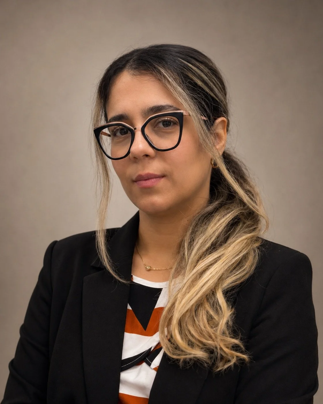Portrait of a woman with long, wavy, blonde hair and glasses, wearing a black blazer and patterned shirt, against a plain background.
