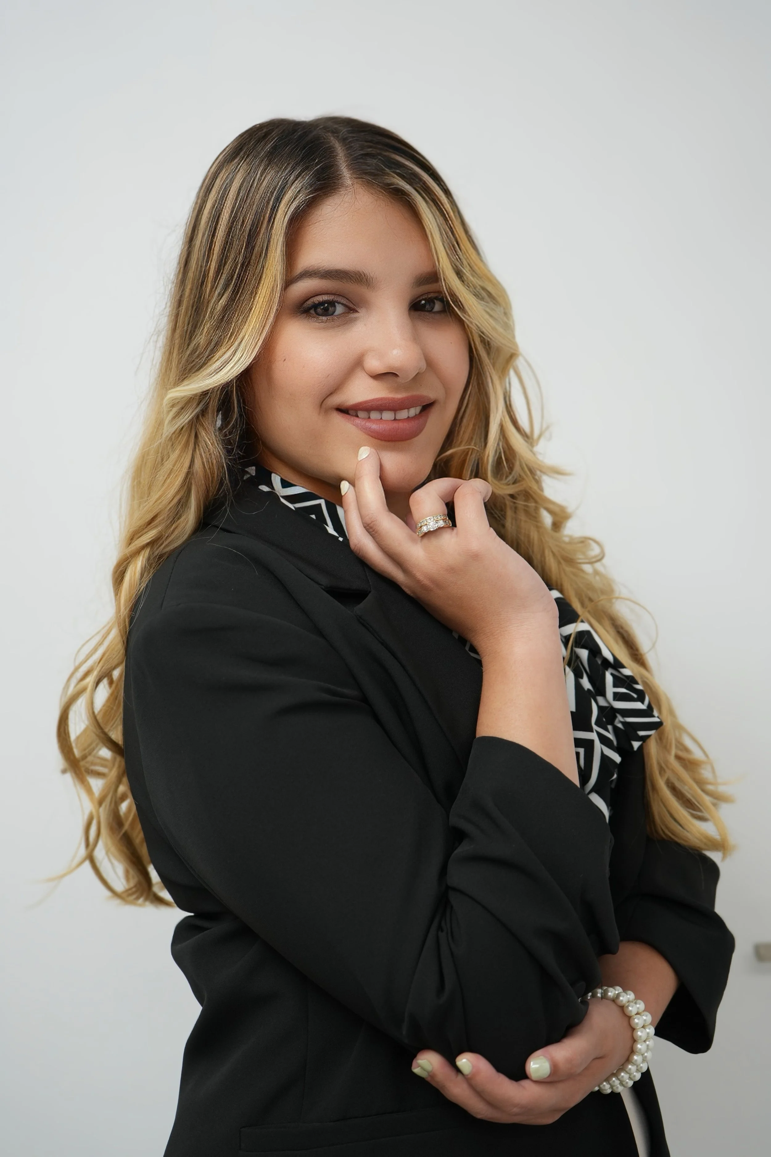 A woman with long, wavy blonde hair wearing a black blazer, pearl bracelet, and rings, posing with a confident smile against a plain white background.