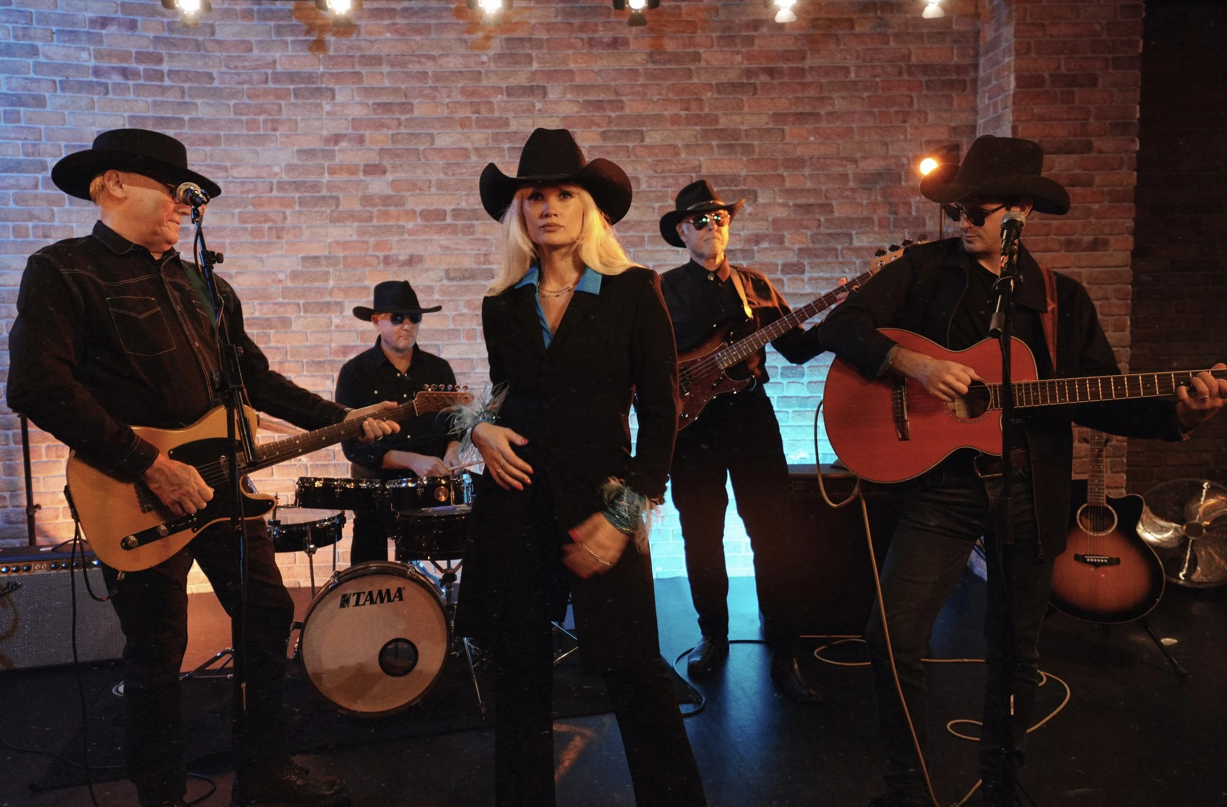 A band performing on stage with a woman singer and four male musicians wearing black cowboy hats, playing guitars and drums against a brick wall background.