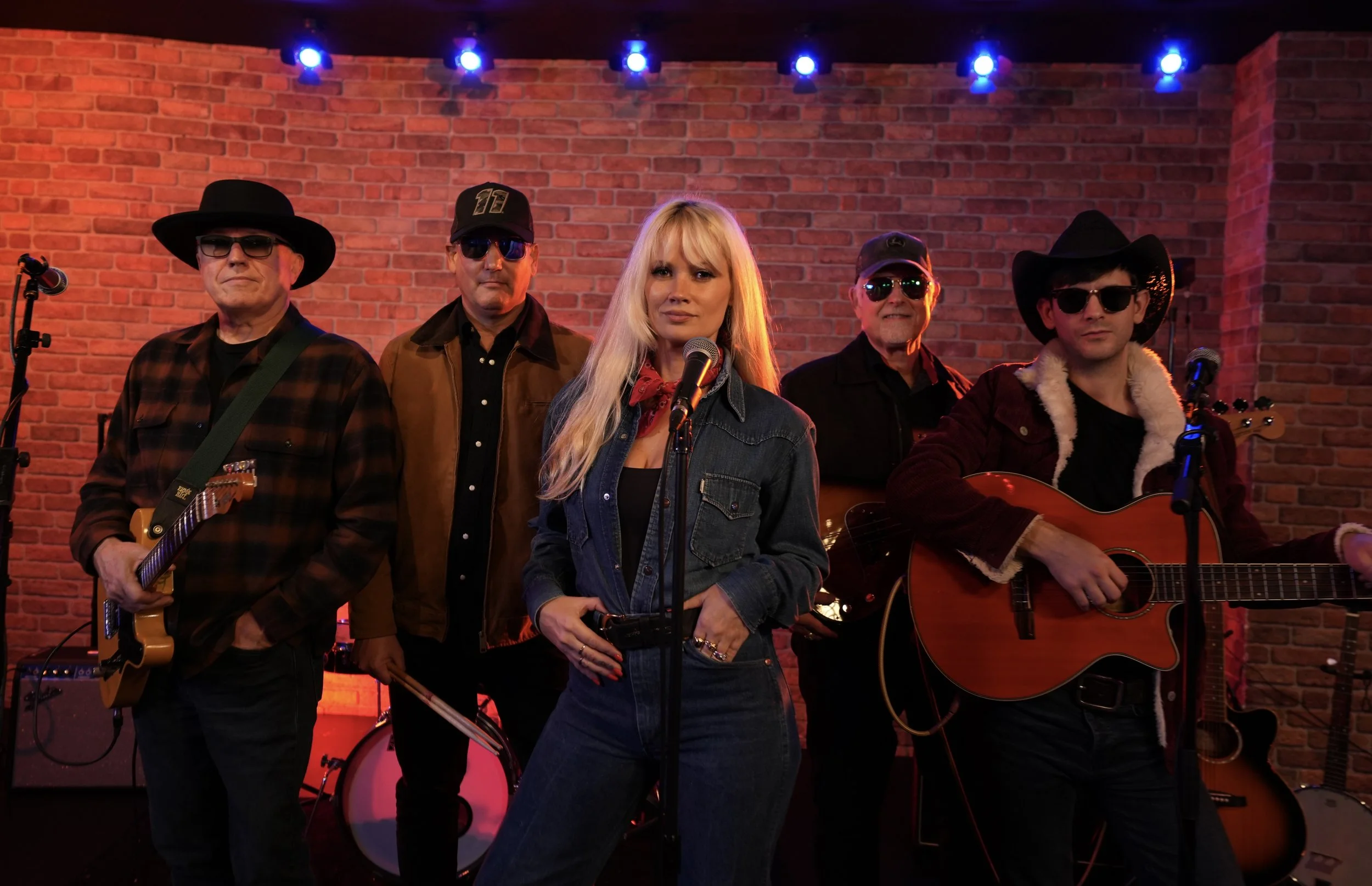 A woman with long blonde hair wearing a denim jacket and red bandana stands in front of a brick wall stage with four men, three with sunglasses and cowboy hats, holding guitars and microphones, under blue stage lights.