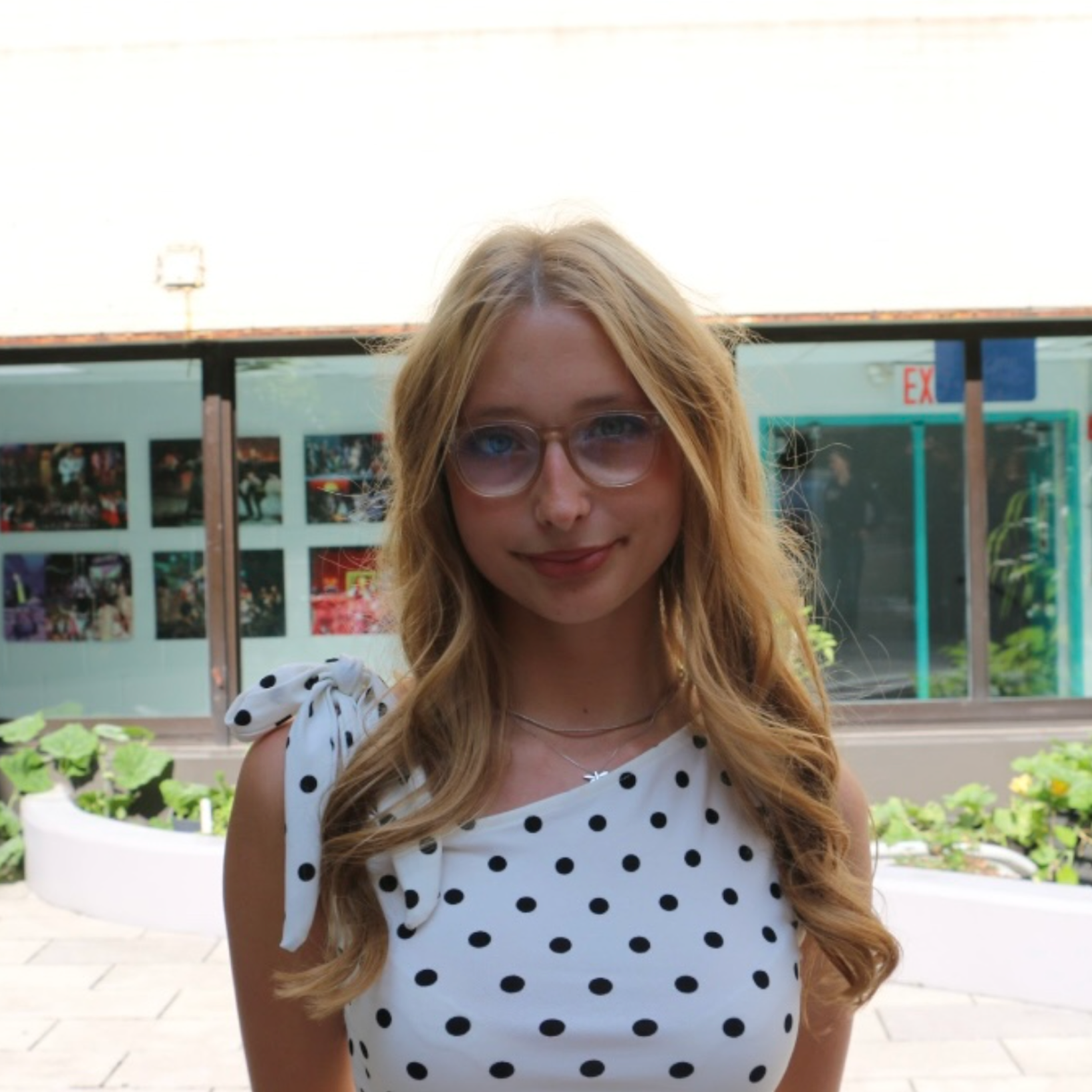 A young woman with long, wavy blonde hair wearing a white dress with black polka dots and glasses, standing outdoors near a window with a display of photos.