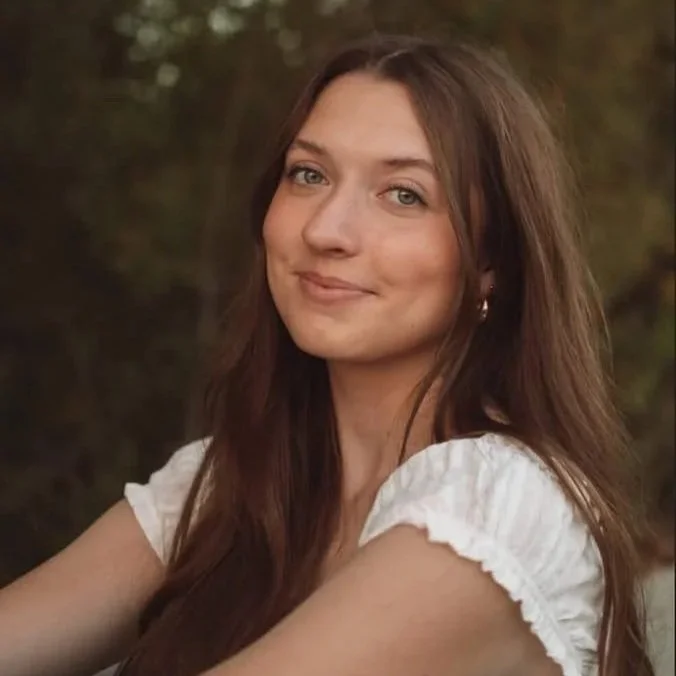 A young woman with long brown hair, standing outdoors with blurred greenery background, smiling softly at the camera, wearing a white top.