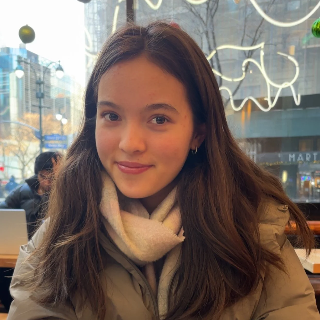 A young woman with long brown hair and hoop earrings sitting inside a cafe, with a city street visible through the window behind her.