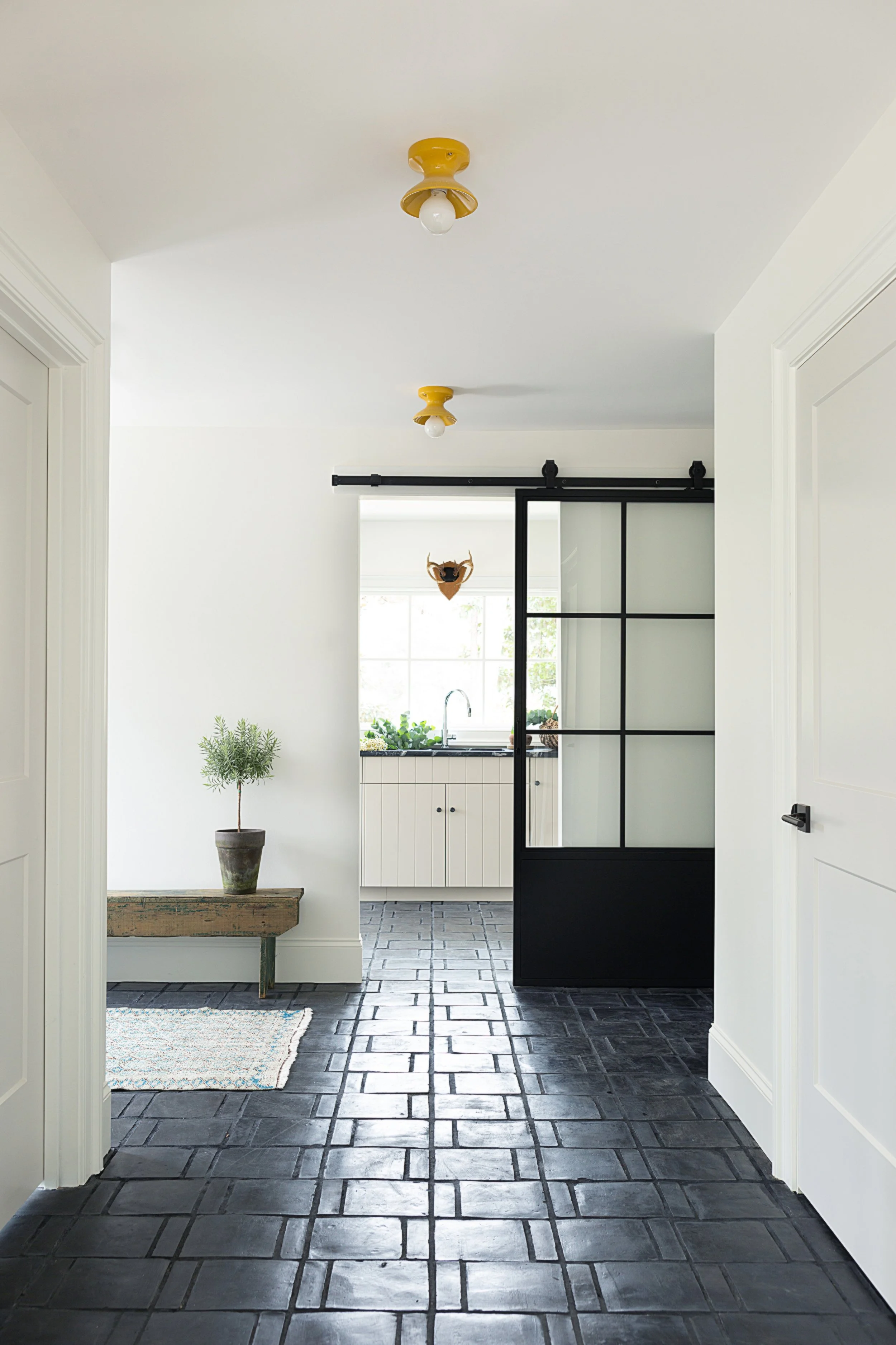Hallway with reclaimed terracotta tile floors, mustard yellow ceramic ceiling lights, and a vintage wood bench with an olive tree, opening through a black steel sliding door to a kitchen with beadboard cabinets