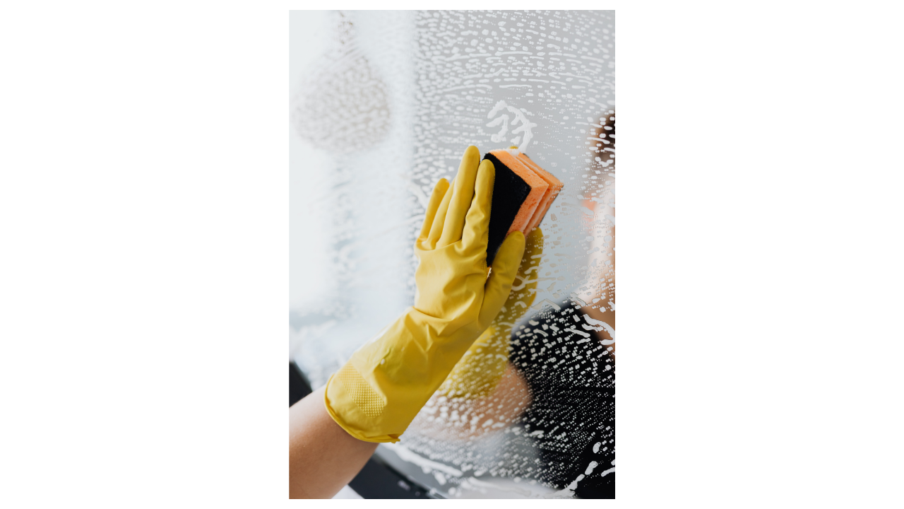 Person wearing yellow rubber gloves cleaning a glass window with a sponge, with soap and water visible.
