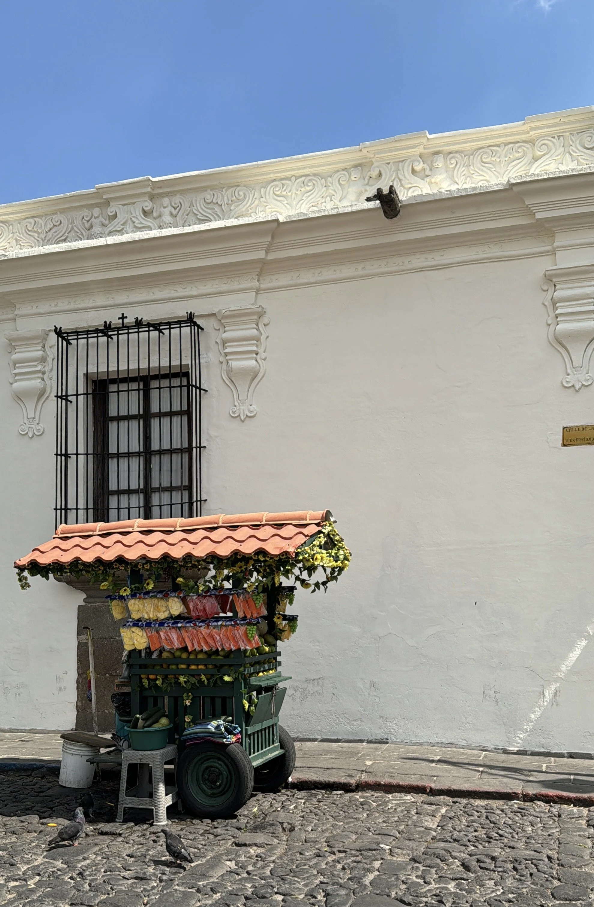 Street vendor cart with a pink-tile roof selling fruits and vegetables, parked on cobblestone street outside a white building with decorative molding and barred window, under a bright blue sky.