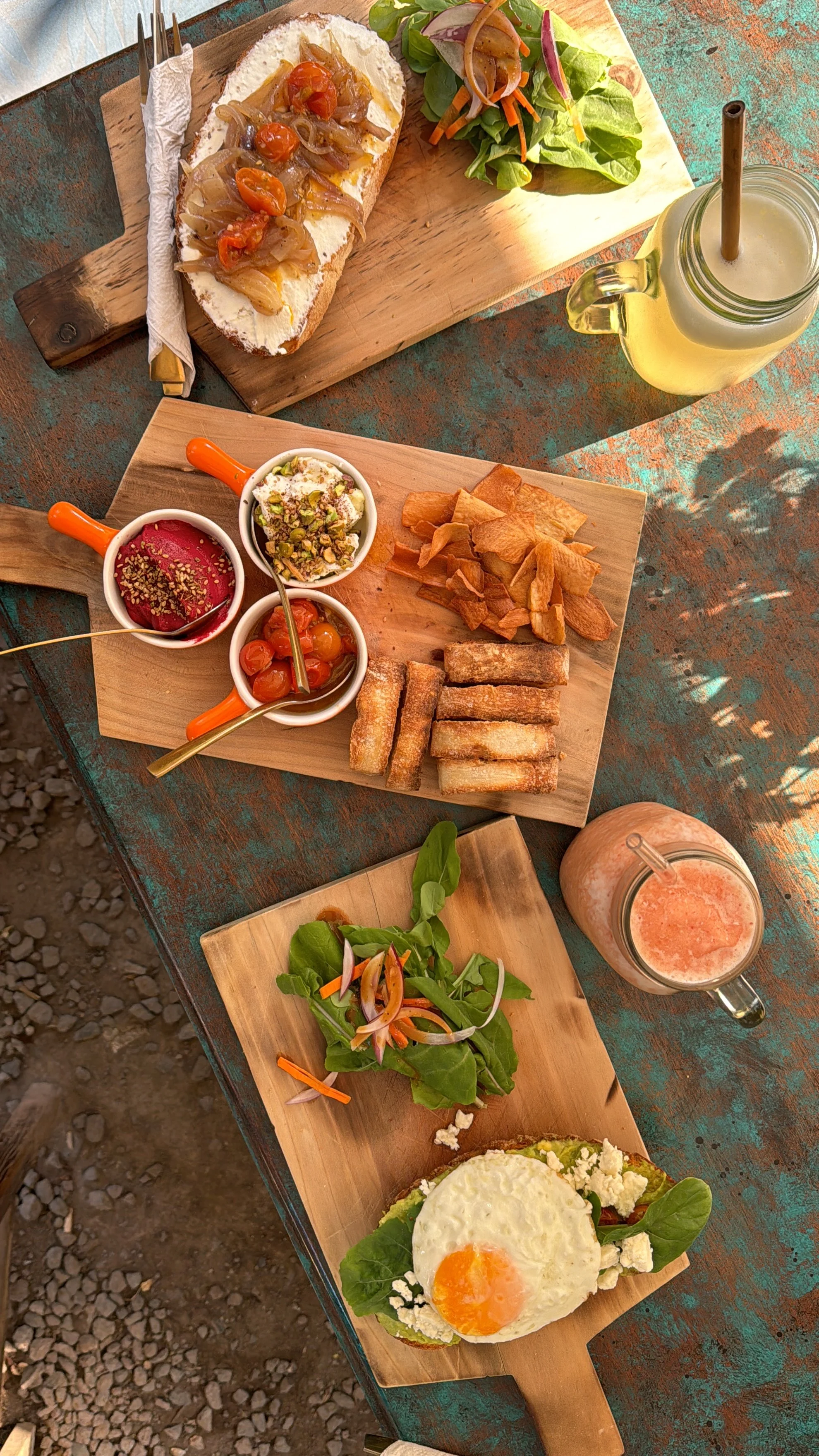 Various breakfast dishes on wooden boards, including eggs with greens and a fried egg on toast, a selection of small bowls with dips and toppings, fresh salads, and beverages such as a yellow drink and a pink smoothie.
