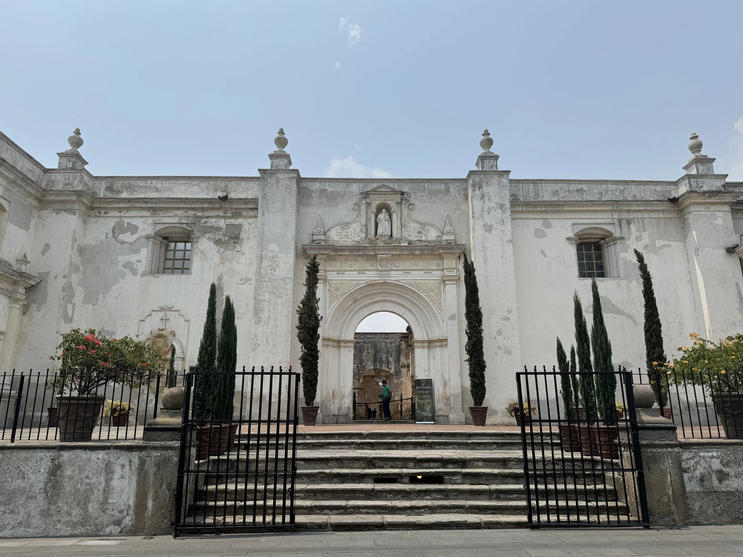 Front view of an old, white, colonial-style building with a central arched entrance, two tall cypress trees on each side, and a few potted plants on the steps leading up to it.