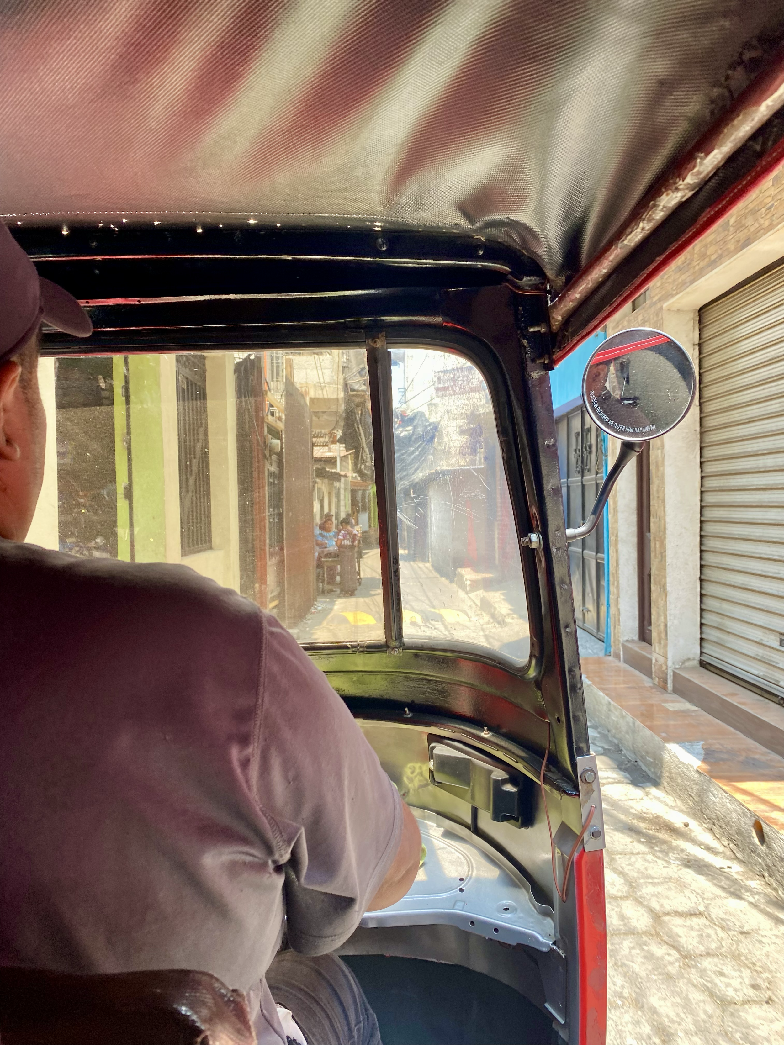 View from inside a tuk-tuk or auto rickshaw showing the driver, with a street scene ahead including buildings, a picket fence, sidewalk, and pedestrians.