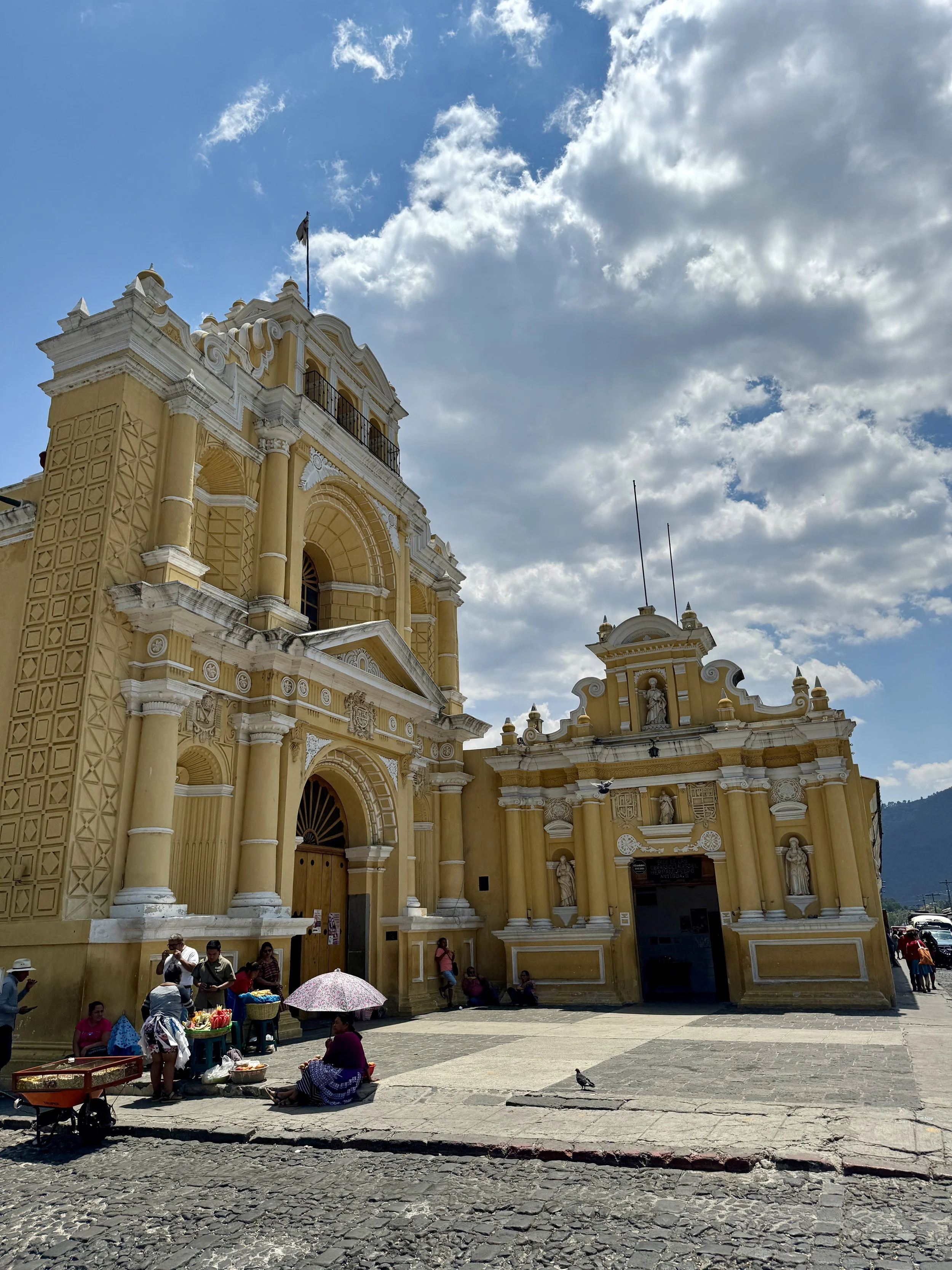 A colonial-style yellow church with white accents and statues in the facade. People are sitting and selling goods outside on a cobblestone street under a partly cloudy sky.