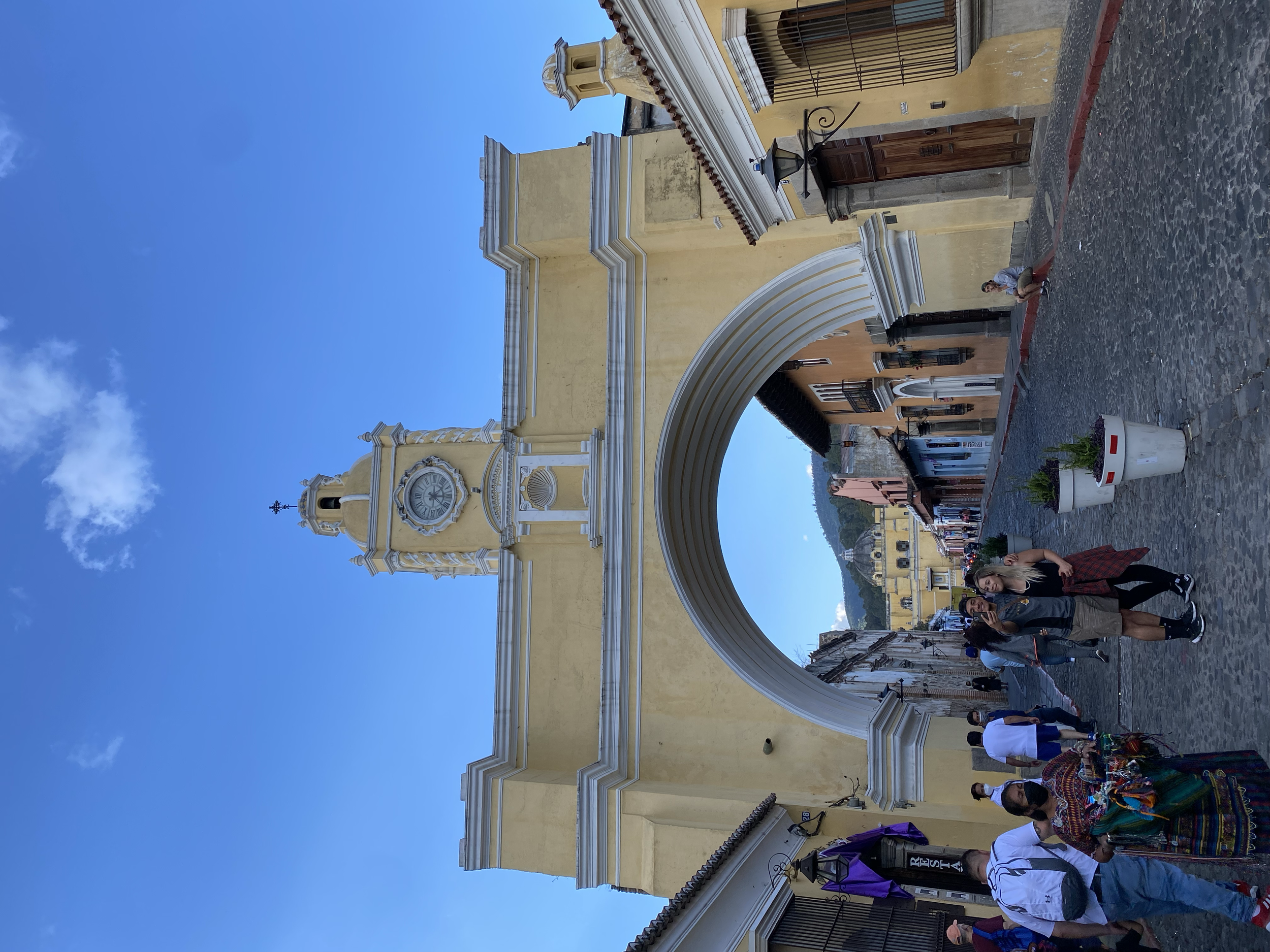 People walking on cobblestone street in front of a yellow church with a clock and bell tower, under a blue sky.