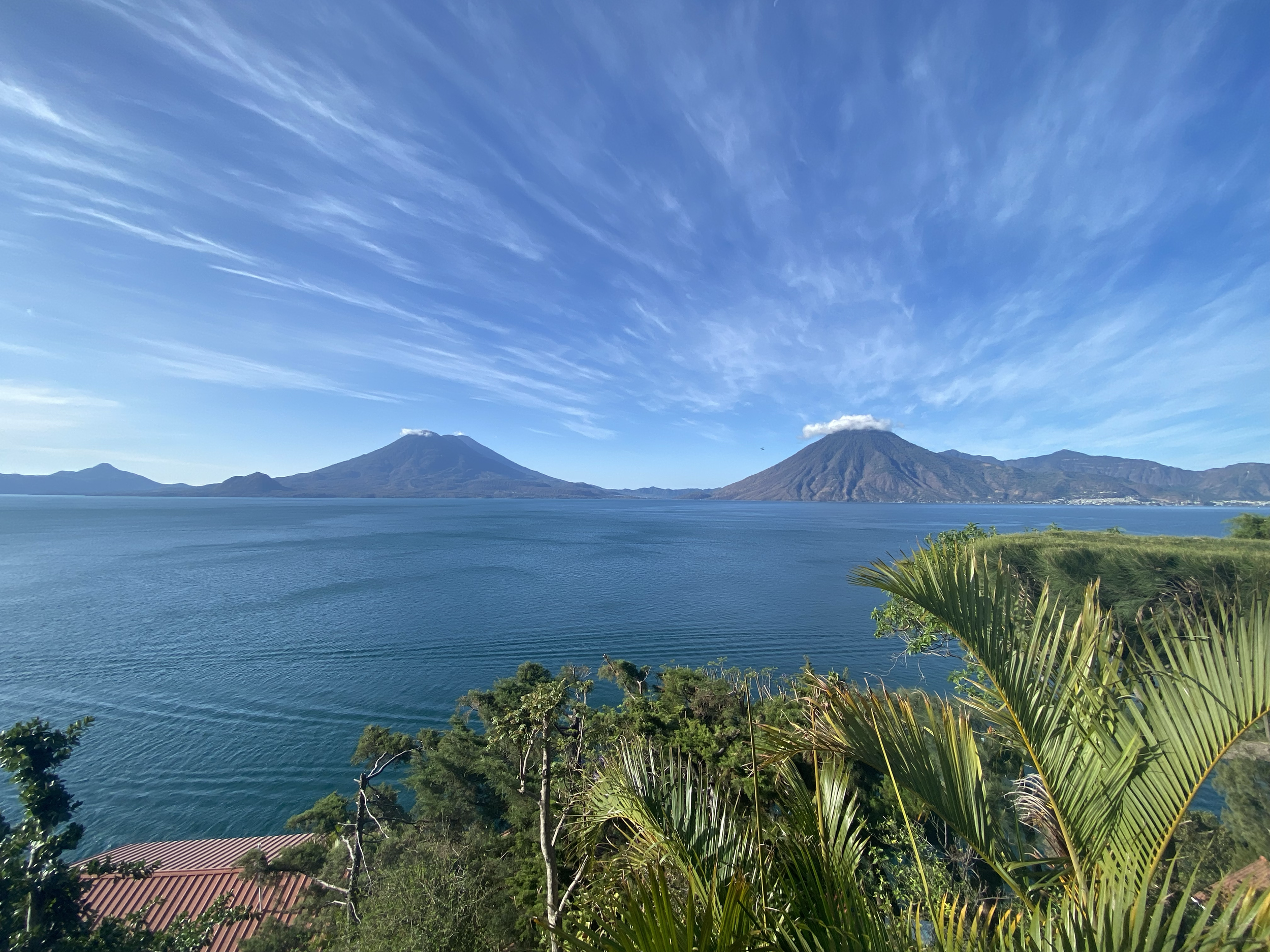A scenic view of a lake with two volcanoes in the background and a blue sky with wispy clouds. Green trees and palm leaves are in the foreground.