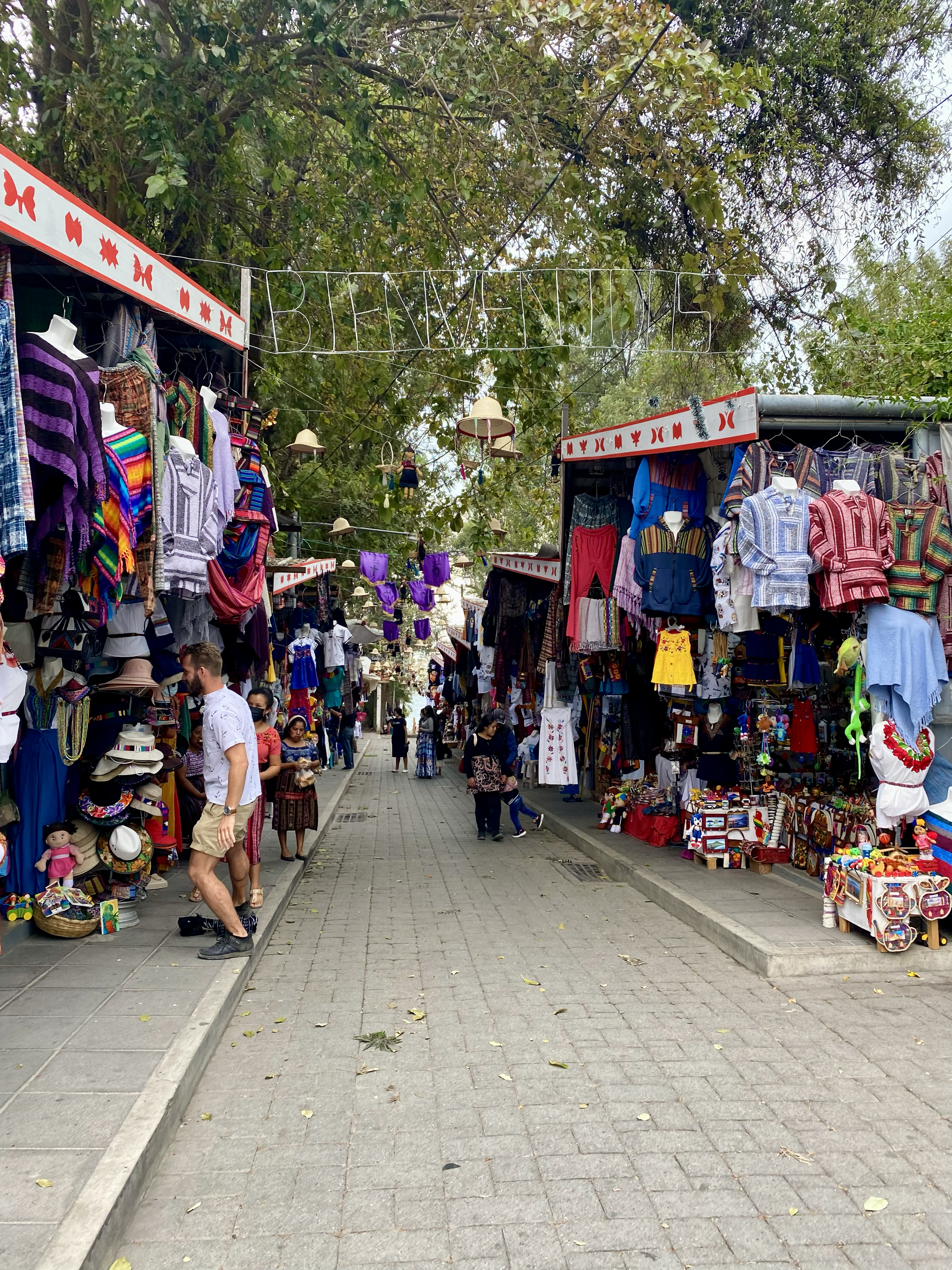 A busy outdoor market with stalls selling colorful clothing, hats, and souvenirs, under tall trees, with shoppers browsing the items.