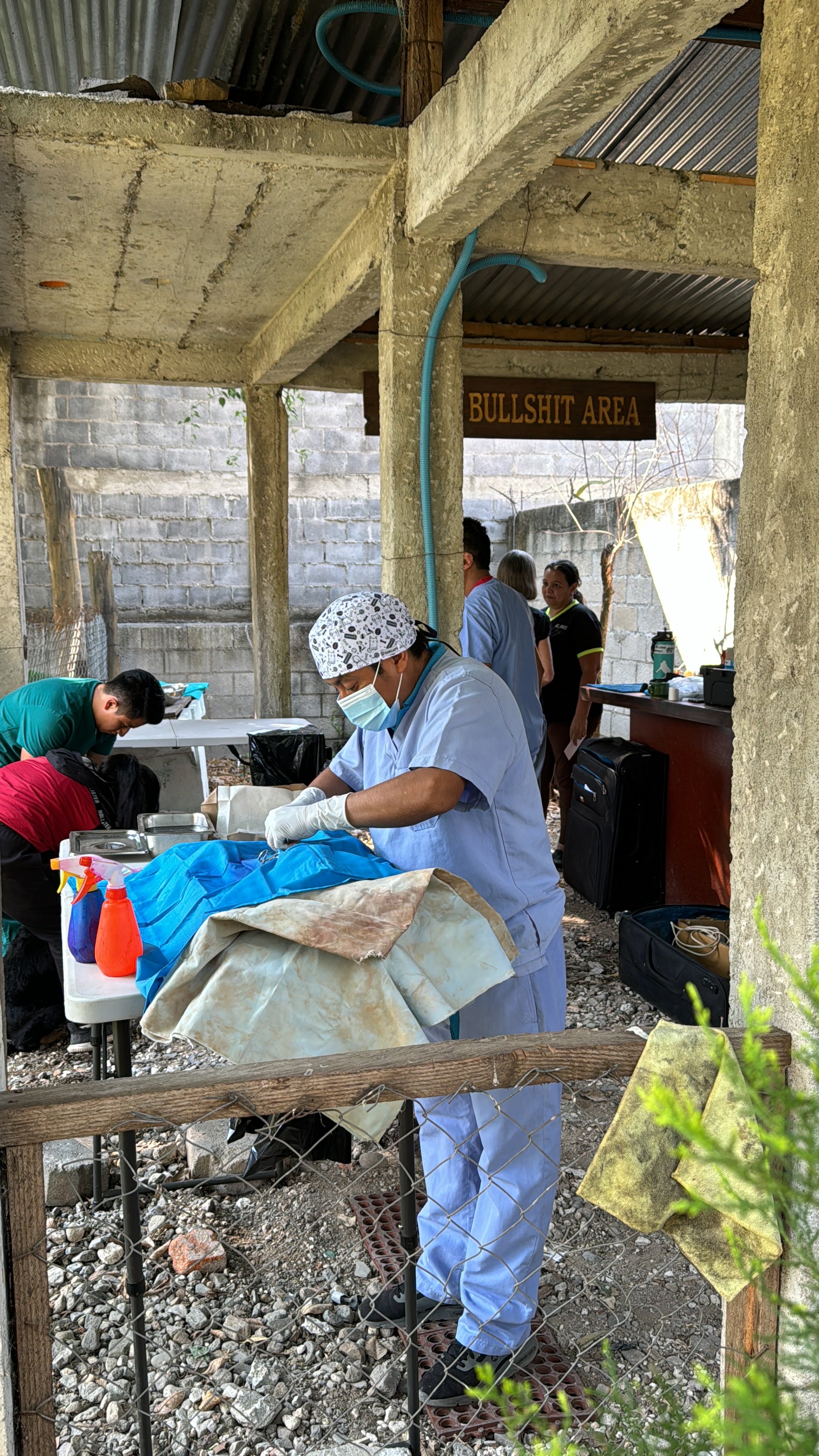Person wearing medical scrubs, gloves, and a face mask performing a procedure outdoors at a medical station labeled None. There are other people in the background, some waiting and some working, in an area marked 'BULLSHIT AREA'. Items such as spray 