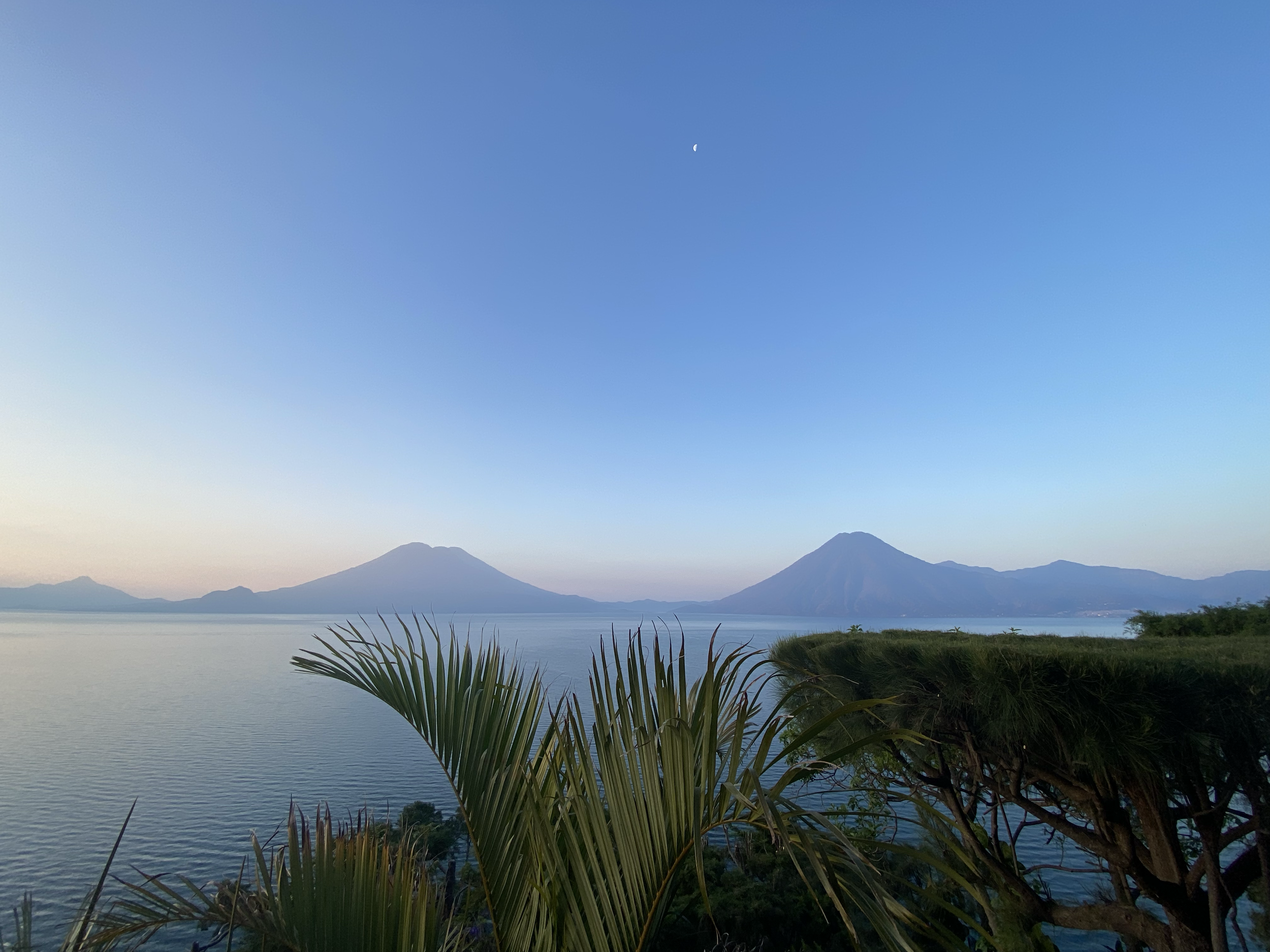 A scenic view of a lake with two volcanoes in the distance under a clear sky with a crescent moon visible.