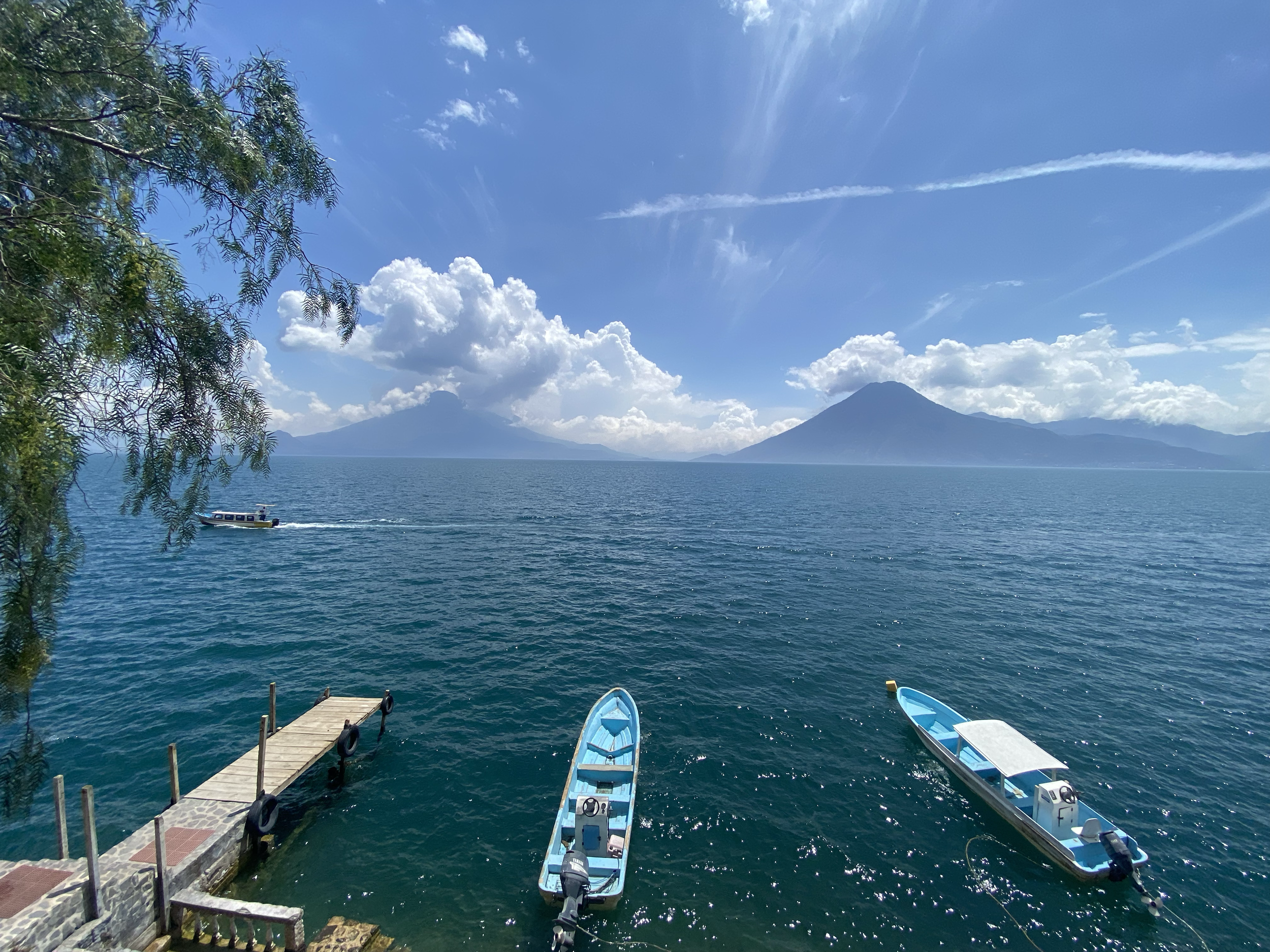 Two small boats are floating on a large body of water near a dock, with mountains and clouds in the background and a partly cloudy sky overhead.