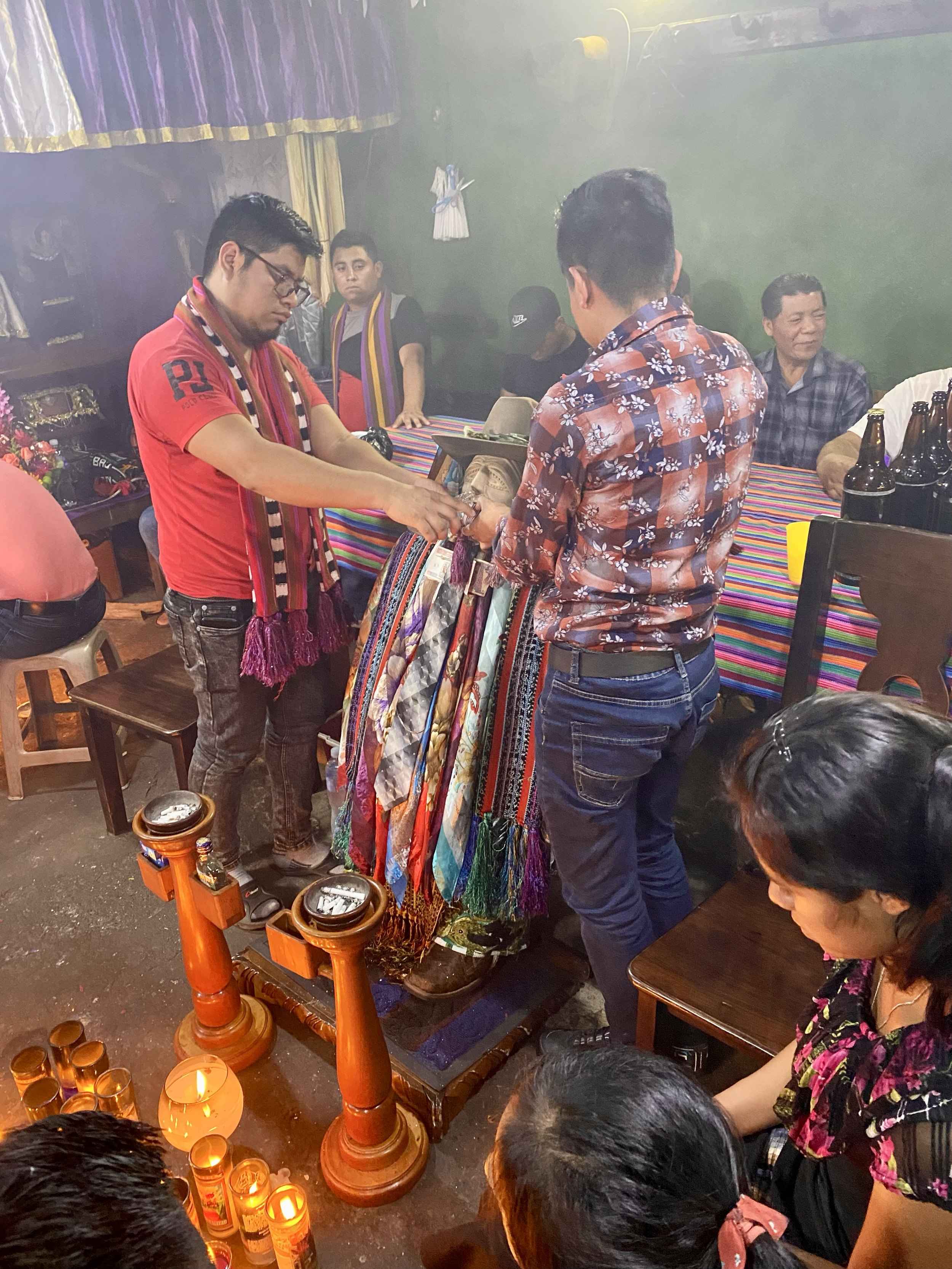 People participating in a traditional ceremony, placing colorful woven textiles on a small platform with candles and incense, in a dimly lit room decorated with traditional fabrics.