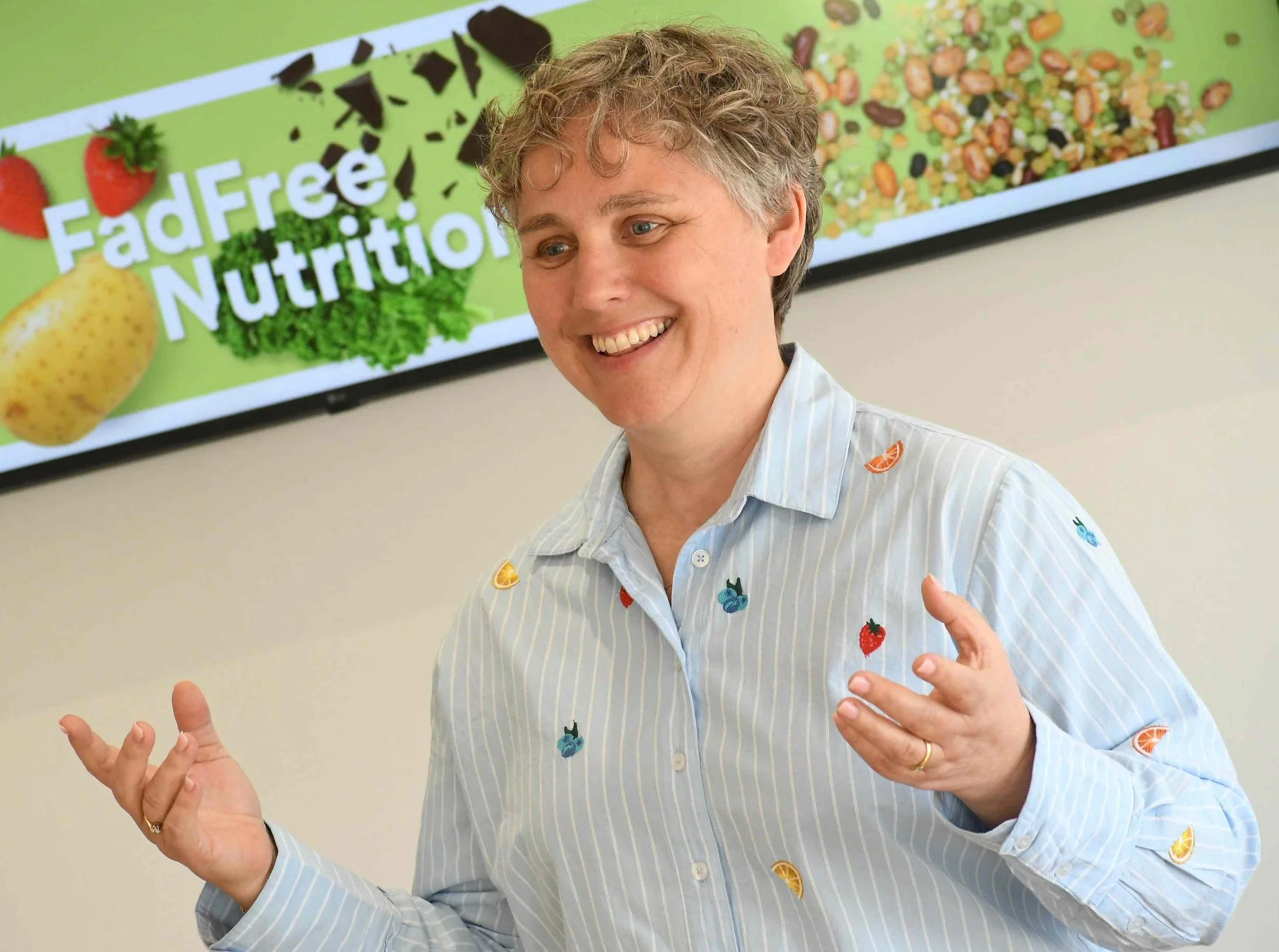 A woman smiling and gesturing with her hands, standing in front of a green sign that says 'Farm to Nutrition' with images of strawberries, an orange, a banana, lettuce, chocolate pieces, and a mixture of beans or seeds.