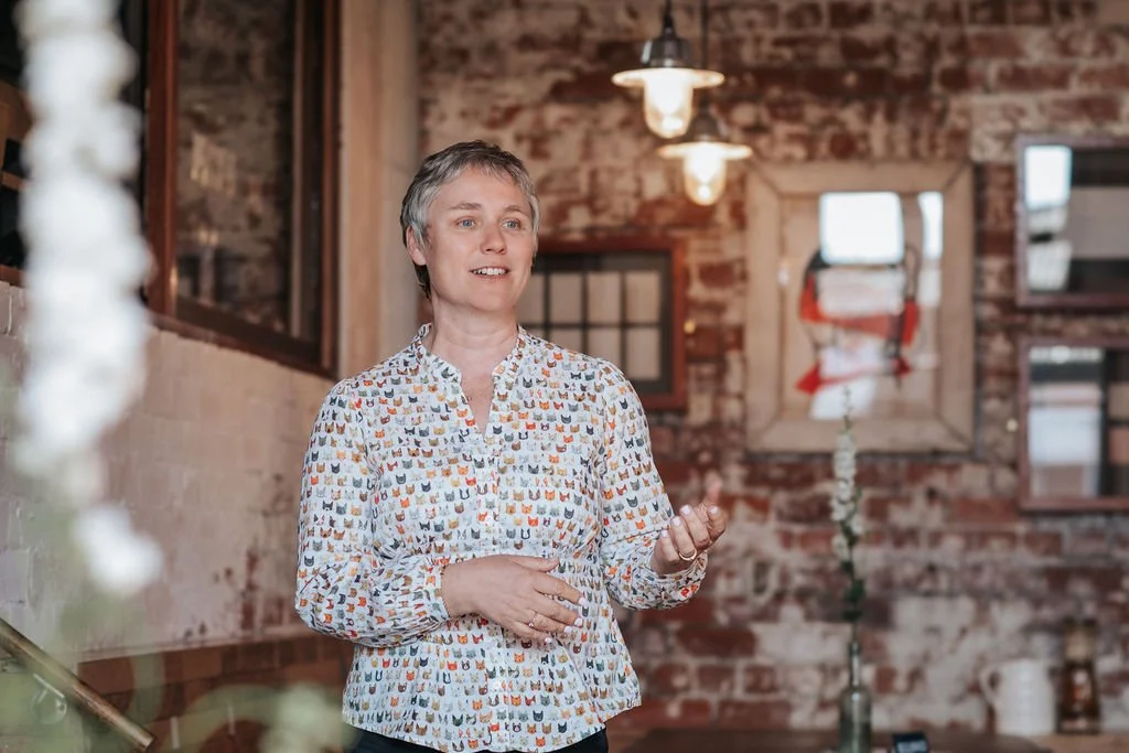 A woman with short gray hair speaking or smiling, wearing a blouse with a colorful cat pattern in a rustic brick-walled room with hanging lights and a window in the background.