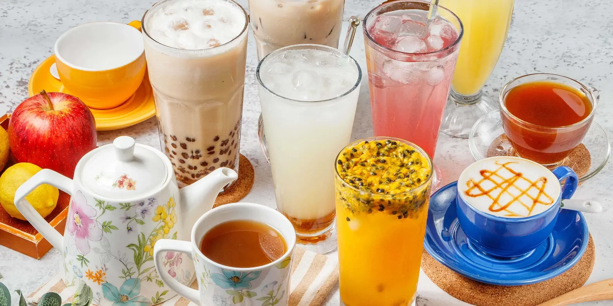 An assortment of breakfast drinks on a table, including bubble tea, fruit juices, tea, and coffee, along with fresh fruits like apple, lemon, and passion fruit.