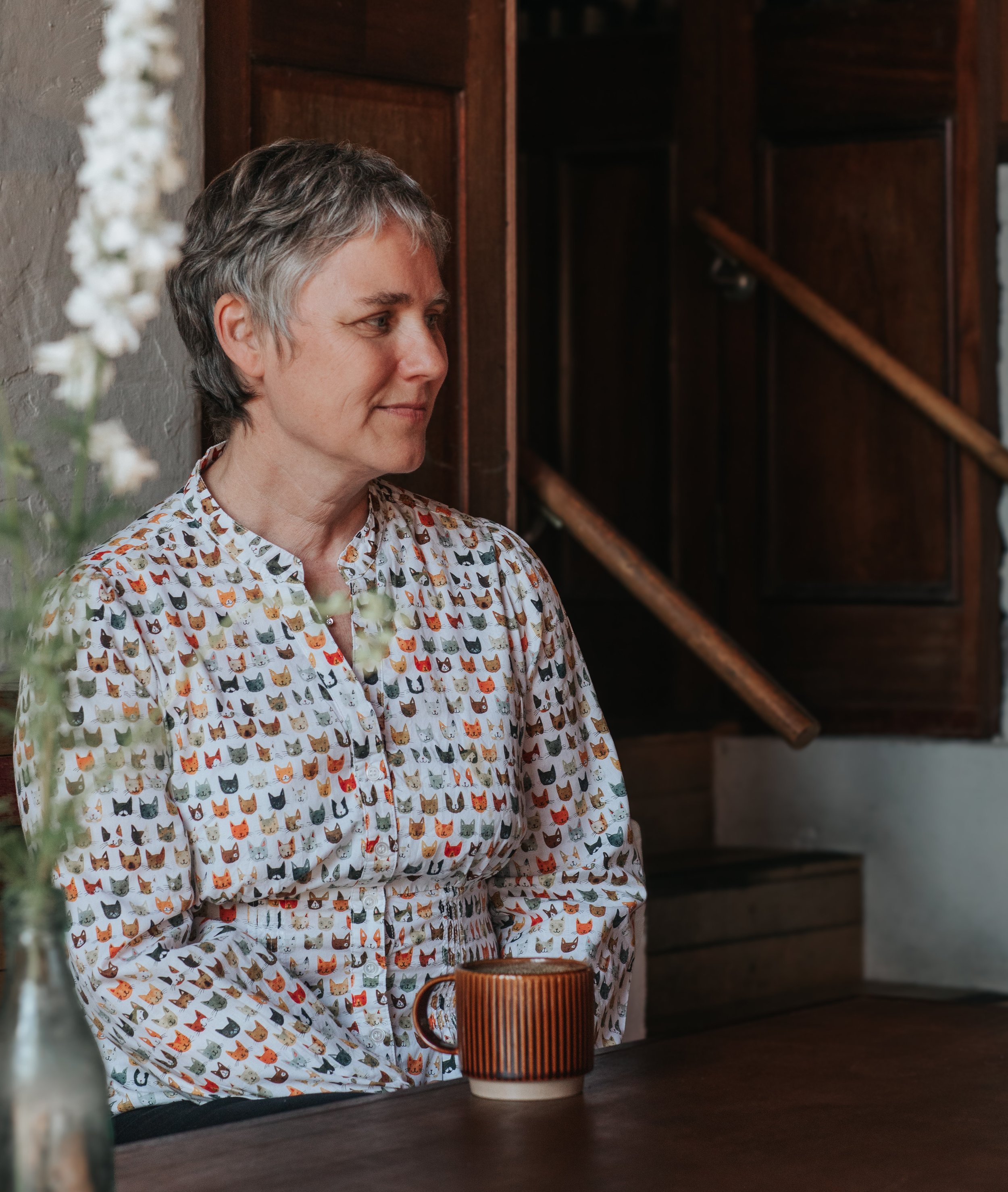 A woman with short gray hair wearing a button-up shirt with a colorful cat print, sitting at a wooden table with a striped mug in front of her, in a cozy room with dark wood paneling and a brick wall.