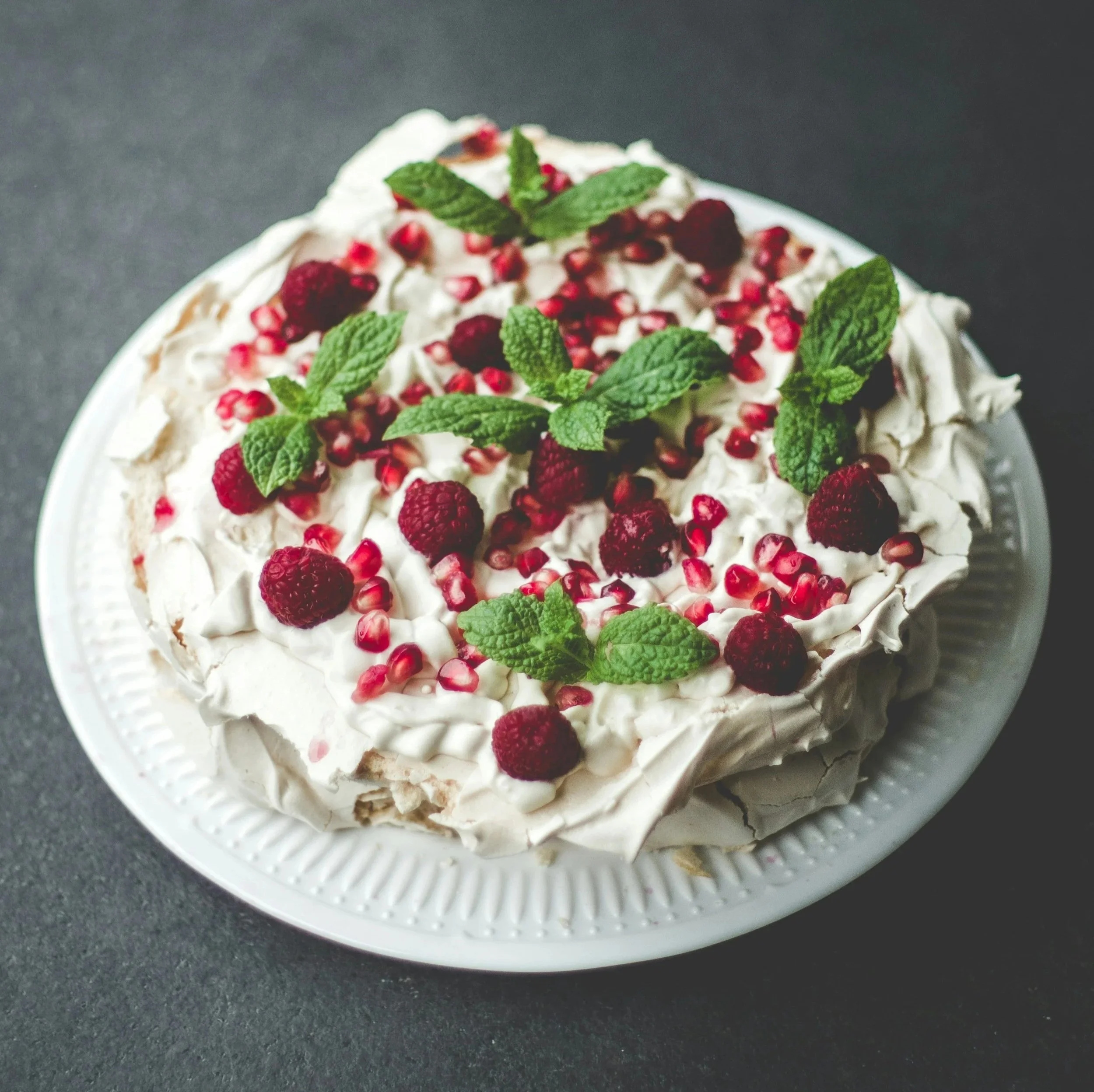 A dessert cake topped with whipped cream, fresh raspberries, pomegranate seeds, and mint leaves on a white plate.