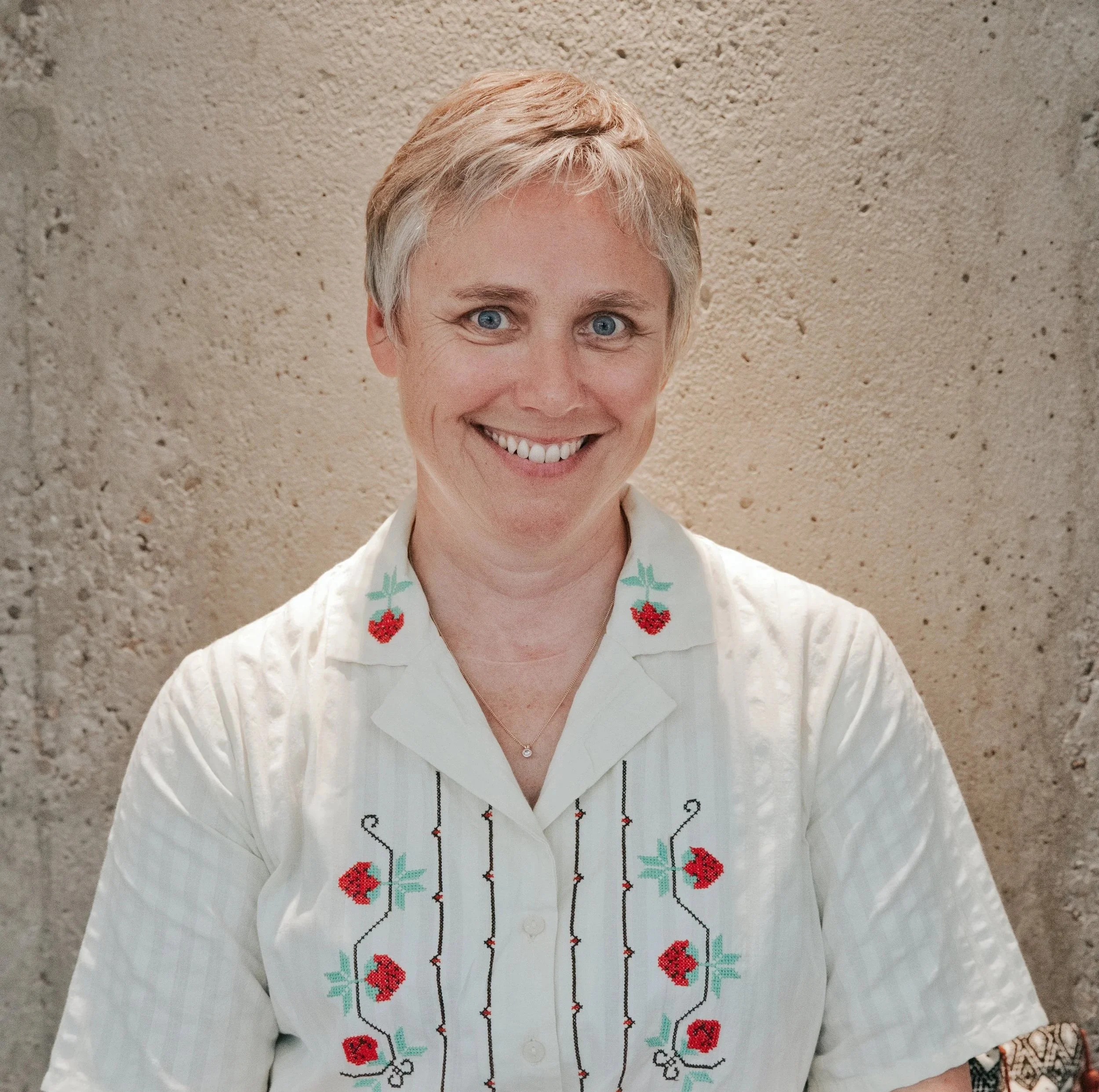 Close-up of a smiling woman with short blonde hair wearing a white blouse with embroidered cherries, standing against a beige stucco wall.
