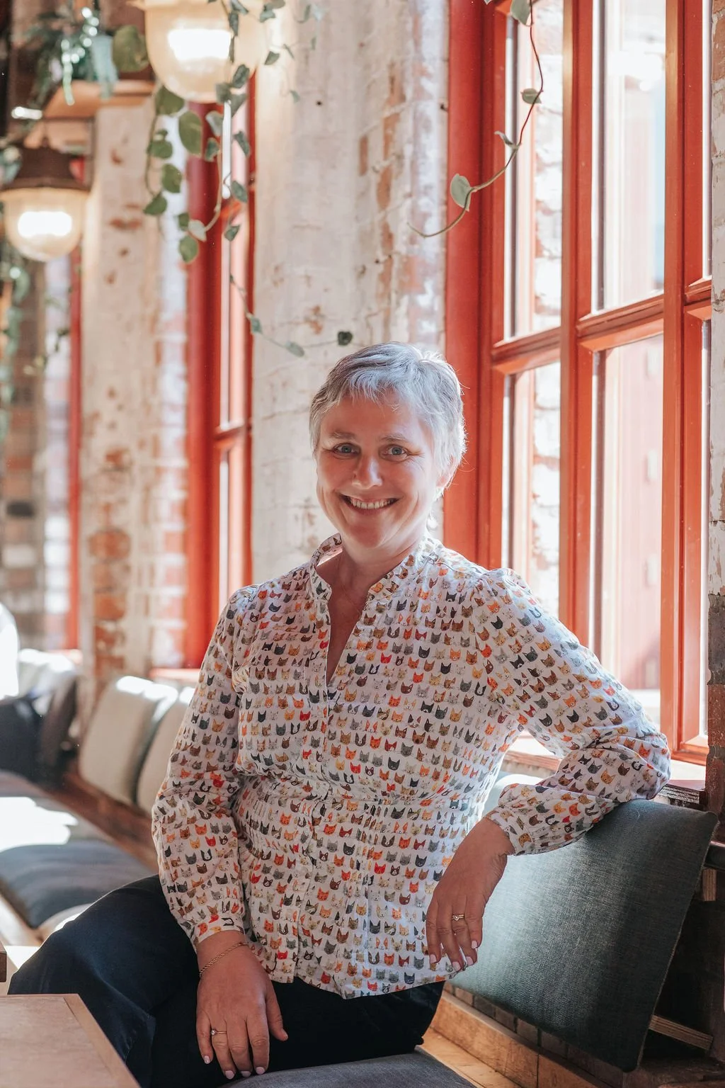 A woman with short gray hair smiling, wearing a button-up shirt with a cat pattern, sitting in a cozy, sunlit room with brick walls and large windows.