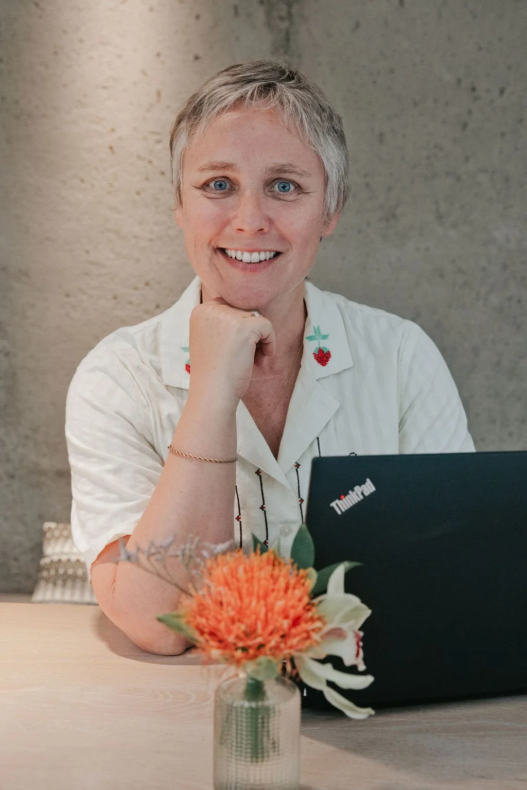 A woman with short gray hair and blue eyes smiling, resting her chin on her hand, sitting at a table with a flower arrangement in a vase and a black Lenovo ThinkPad laptop in front of her. She wears a white shirt with embroidered cherries on the collar.