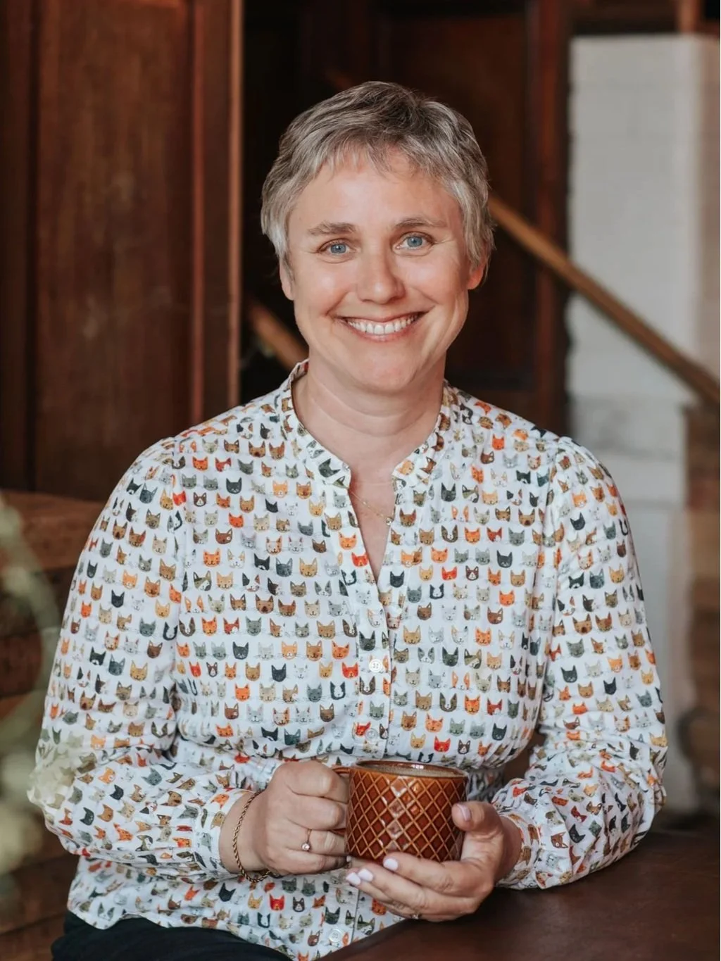 A woman with short gray hair, smiling, holding a brown patterned mug, wearing a white blouse with a colorful cat face print, standing indoors in a cozy setting.