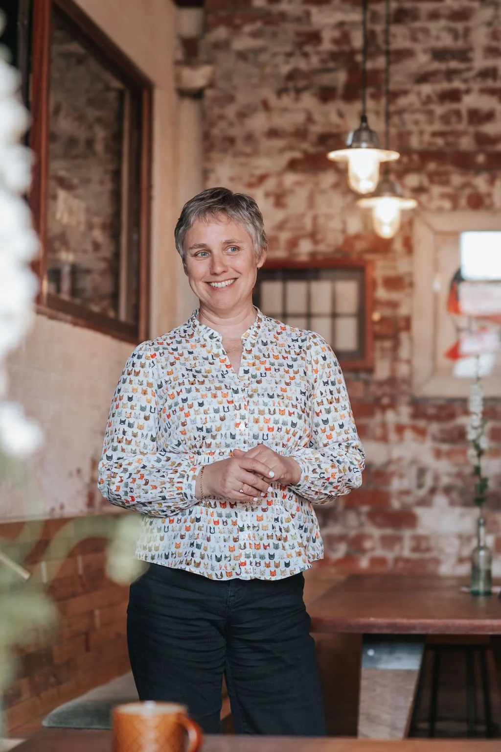 A woman with short hair smiling, standing indoors with a brick wall background, wearing a button-up shirt with a colorful cat pattern.