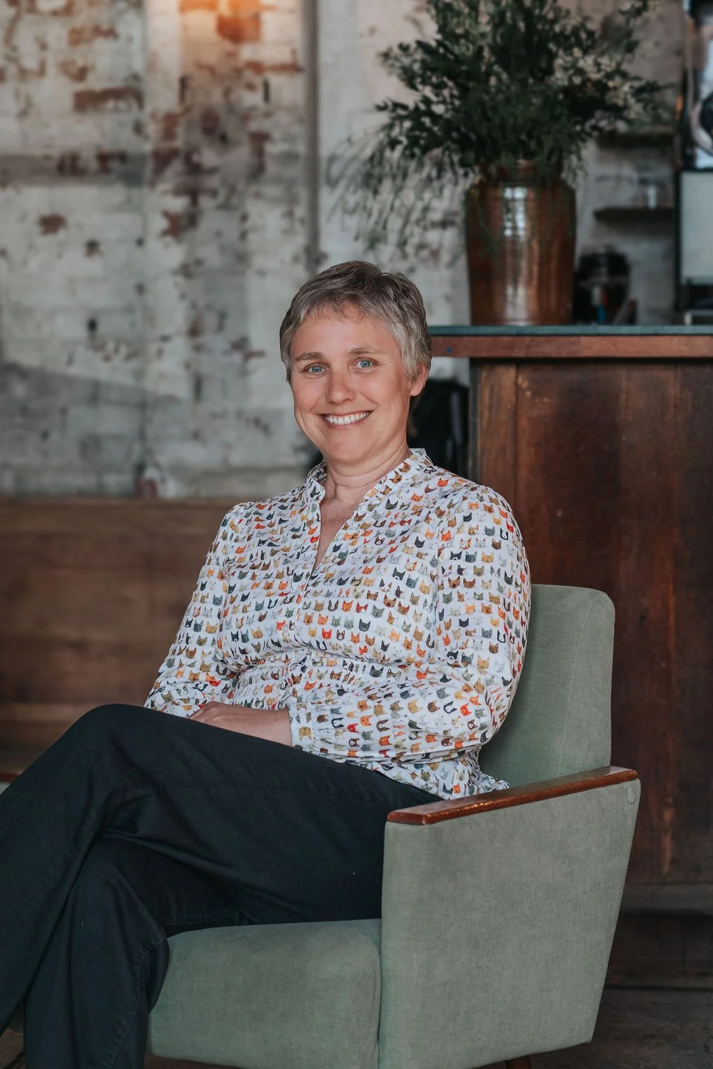 A woman sitting in a green armchair, smiling, with a brick wall and a plant in the background.
