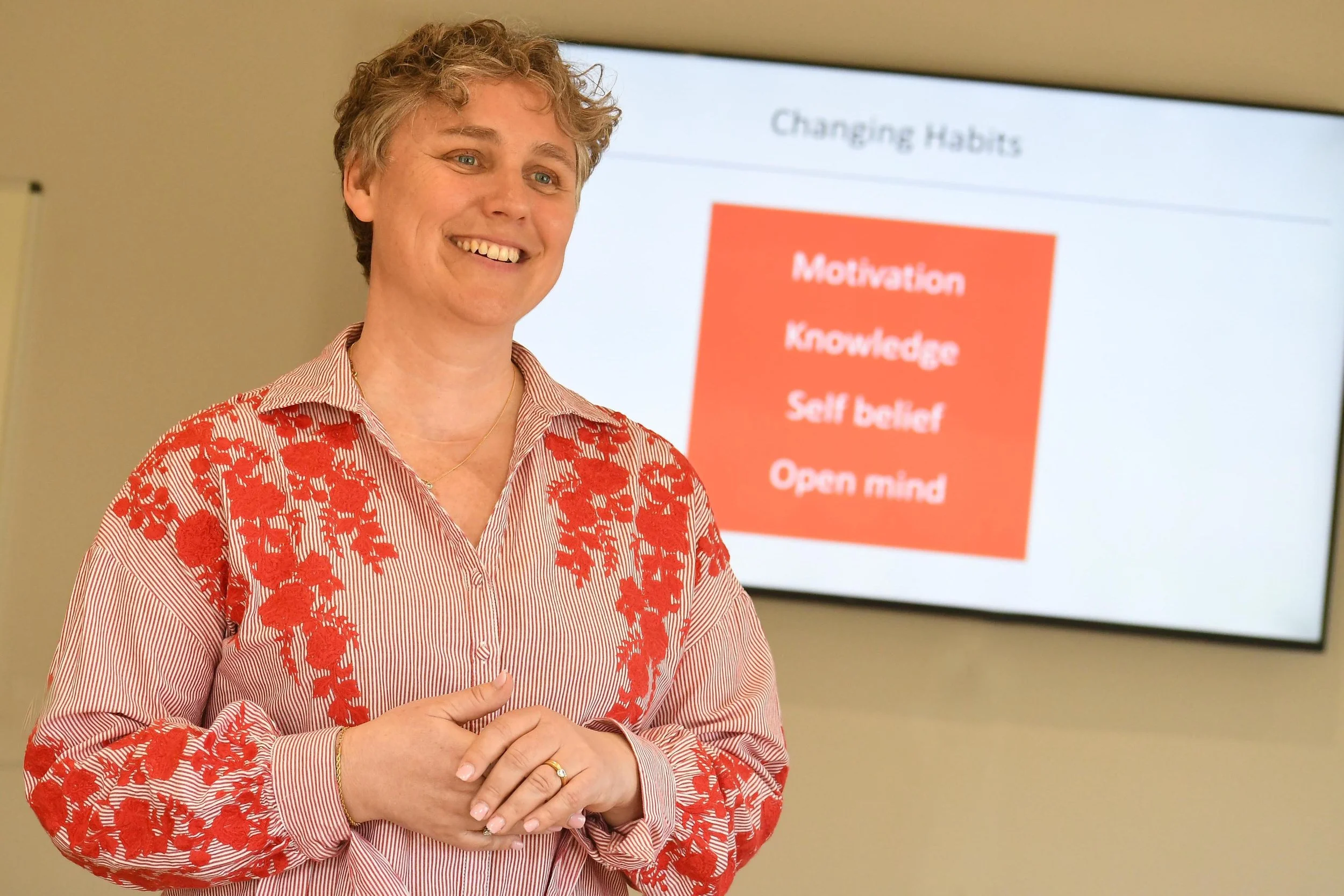 A woman smiling and standing in front of a presentation slide on changing habits, with words like motivation, knowledge, self-belief, and open mind, wearing a red and white floral embroidered shirt.