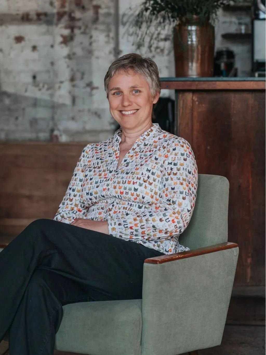 A woman with short hair sitting in a green armchair, smiling. She is wearing a white shirt with a pattern of small colorful cats. Behind her, there is a wooden counter and a large potted plant in a brown pot, with a brick wall in the background.