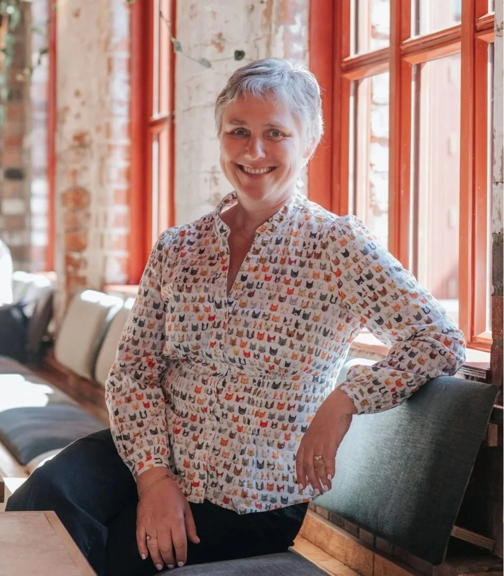 A woman with short gray hair, smiling, sitting on a bench in a room with large windows and exposed brick walls, wearing a colorful cat print blouse.