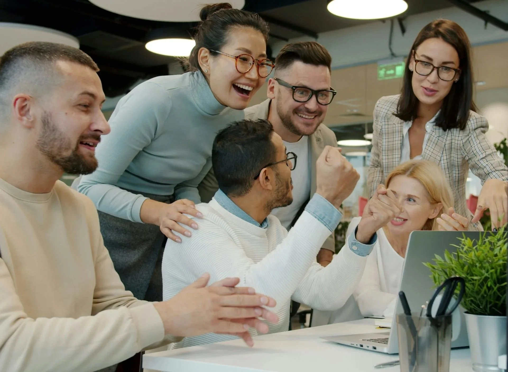 Group of diverse coworkers gathered around a laptop in a modern office, celebrating and smiling.