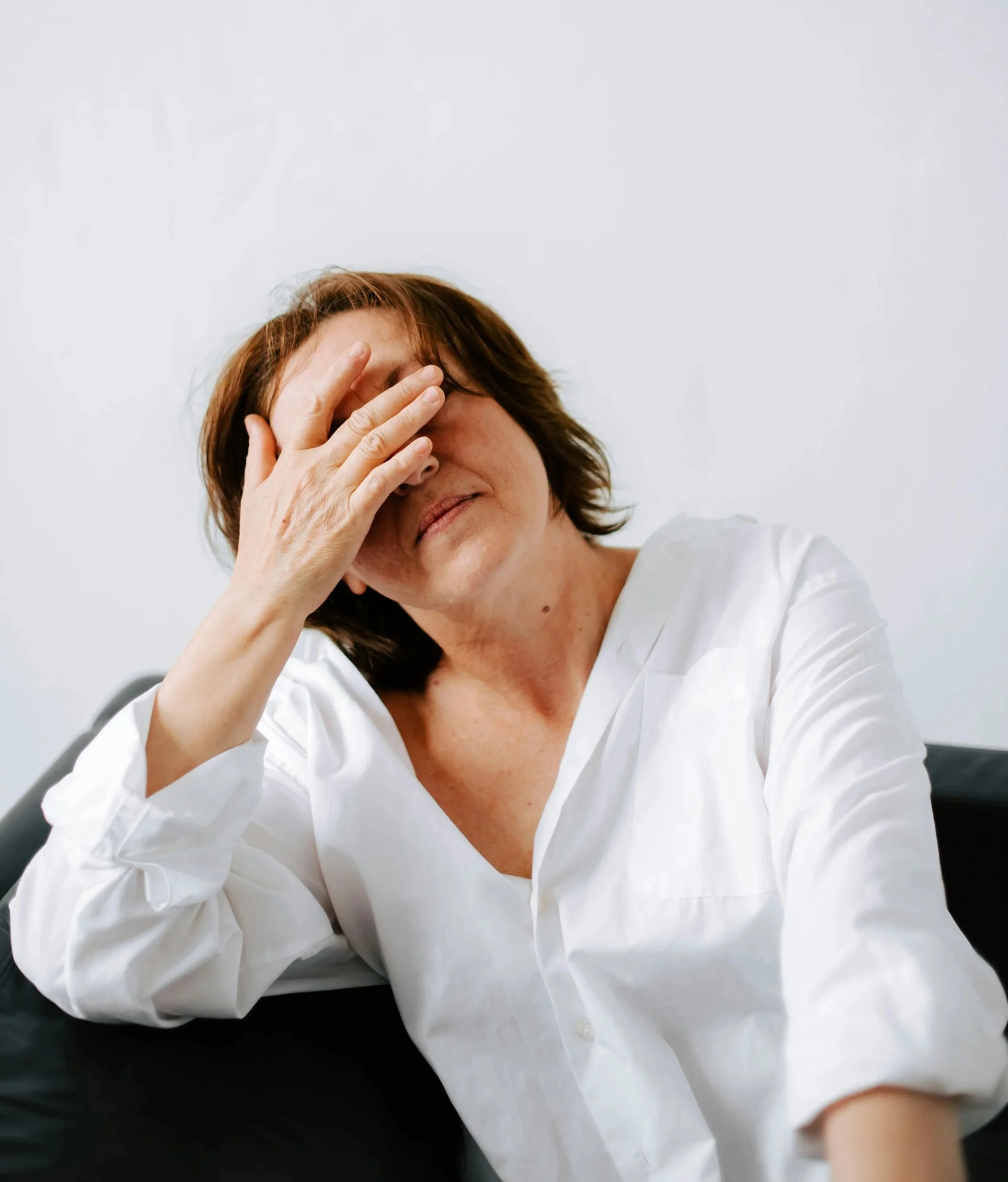A woman with short reddish-brown hair wearing a white shirt, sitting with her hand covering her face, against a plain white background.