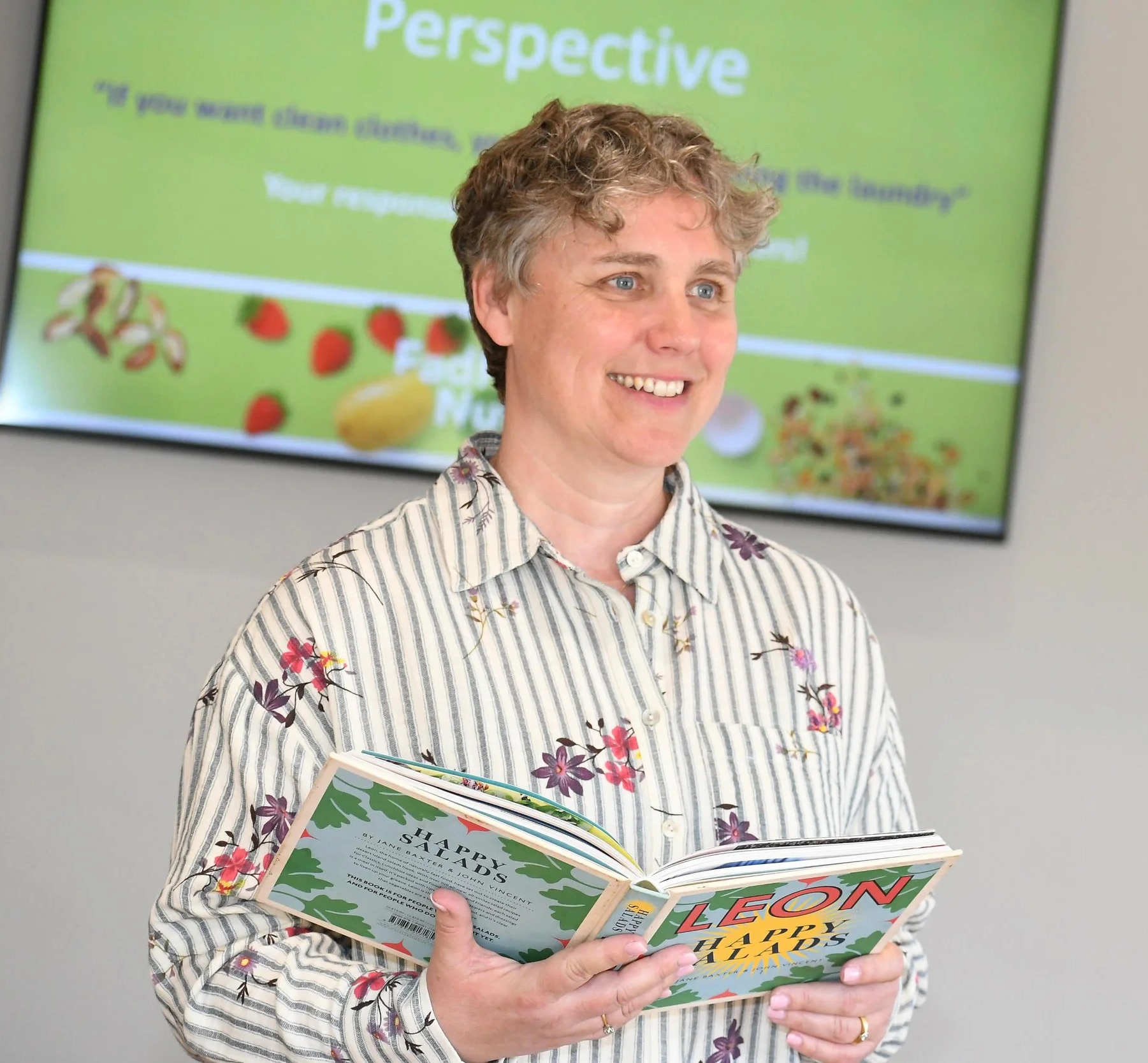 A smiling woman with short curly hair wearing a striped floral shirt holding a children's book titled 'LEON, HAPPY HARDS' in front of a green presentation screen.