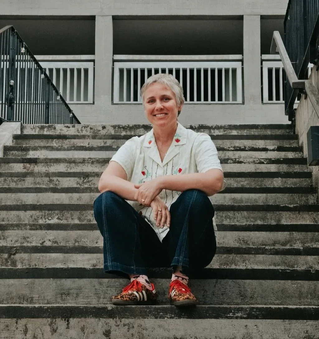 A woman with short blonde hair sitting on concrete stairs, smiling, wearing a white short-sleeved shirt with embroidered cherries, dark jeans, and colorful sneakers, with a building and staircase railing in the background.