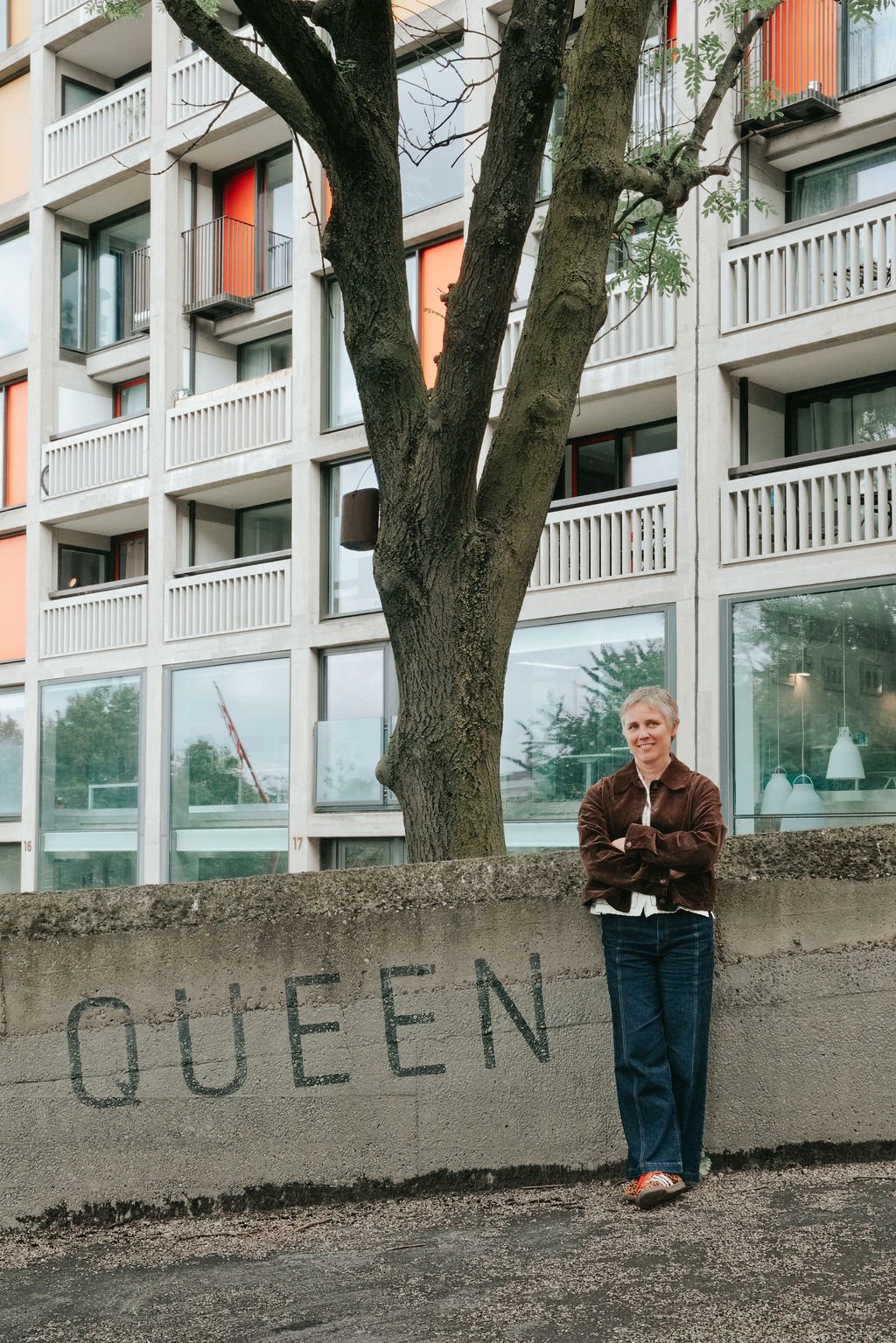 A woman standing with arms crossed against a concrete wall with the word "QUEEN" spray-painted on it. Behind her is a large tree and a modern apartment building with glass windows and balconies.