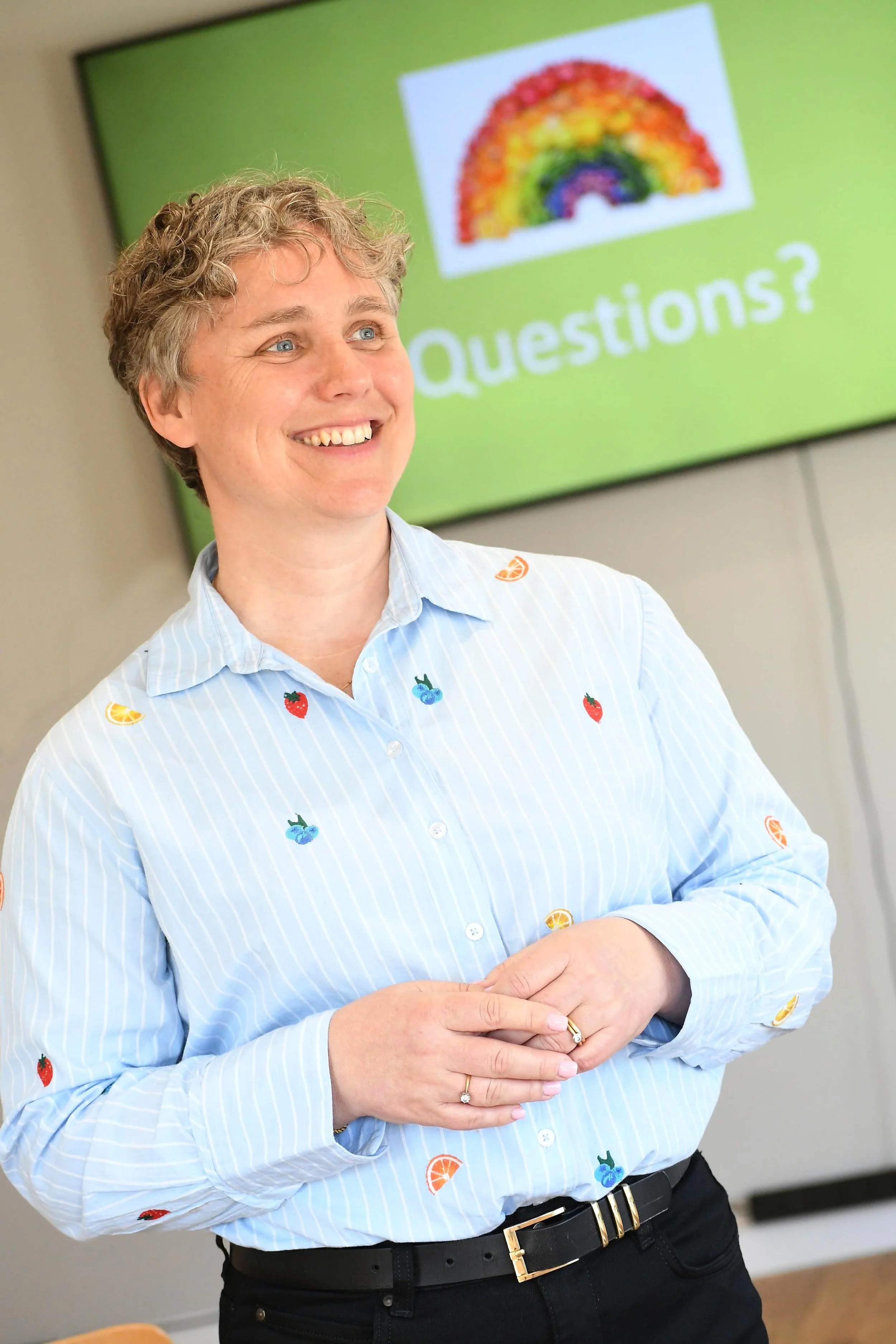 A smiling woman with short blonde hair wearing a light blue shirt with colorful fruit patches, standing in front of a green screen displaying a rainbow drawing and the word 'Questions?'