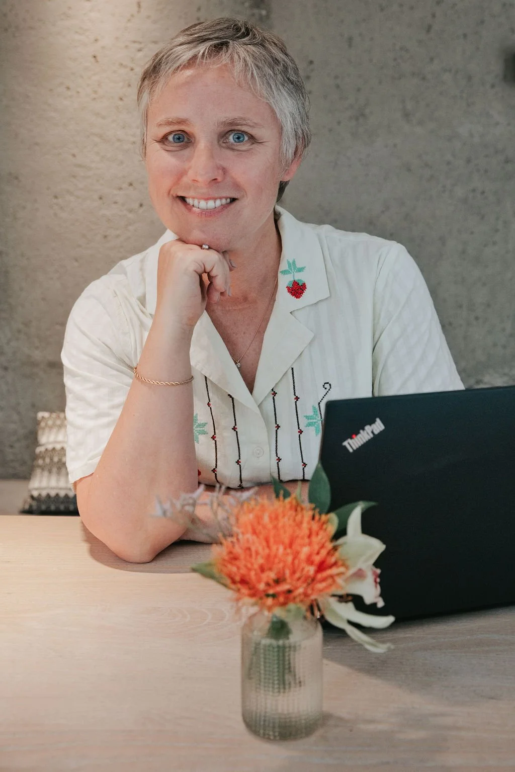 A woman with short gray hair and blue eyes sitting at a desk with a smile, resting her chin on her hand, in front of a laptop and a vase of orange flowers.