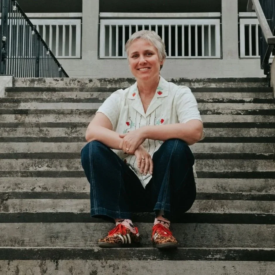 A woman with short gray hair sitting on outdoor concrete stairs, smiling at the camera. She is wearing a light-colored shirt with embroidered flowers, dark jeans, and colorful sneakers with animal print and red laces.